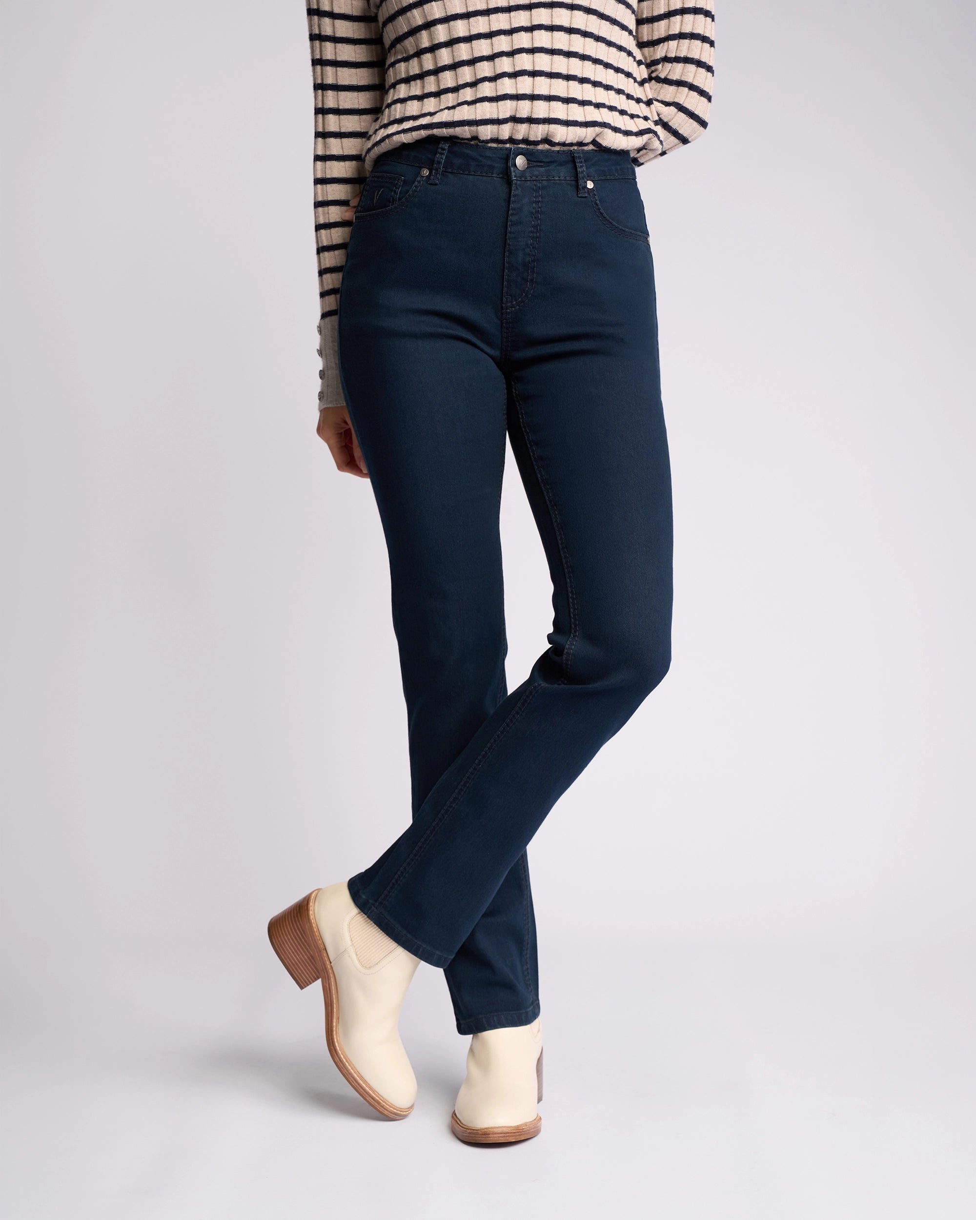 Wearing Vassalli's Lotus Jean in Indigo Denim, paired with a striped long-sleeve top and light ankle boots, a person stands against a plain light background with legs crossed at the ankles.