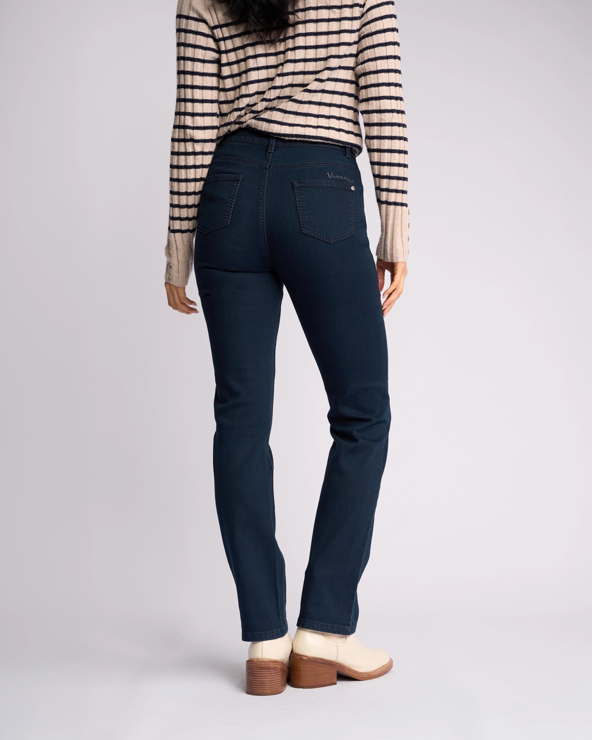 A person stands with their back to the camera, wearing a beige and black striped long-sleeve top, Vassalli Lotus Jean in Indigo Denim, and cream ankle boots with a wooden heel against a plain light background.