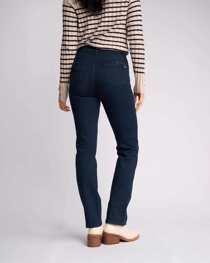 A person stands with their back to the camera, wearing a beige and black striped long-sleeve top, Vassalli Lotus Jean in Indigo Denim, and cream ankle boots with a wooden heel against a plain light background.
