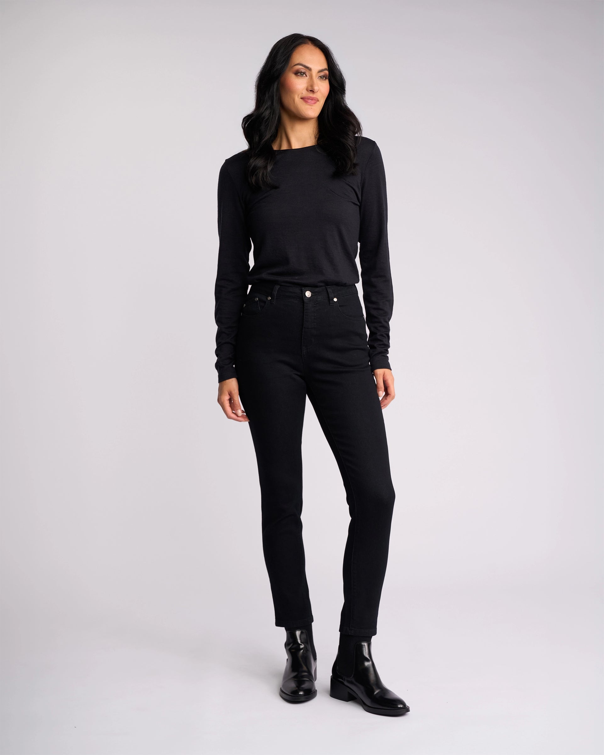 A woman with long dark hair stands against a light gray background, wearing Vassalli’s Icon Jean in Black Denim and a black top. She looks to the side and smiles softly, pairing her outfit with black ankle boots.