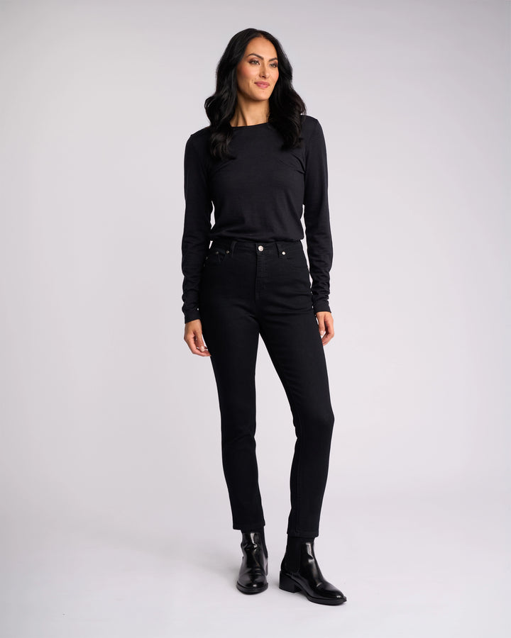A woman with long dark hair stands against a light gray background, wearing Vassalli’s Icon Jean in Black Denim and a black top. She looks to the side and smiles softly, pairing her outfit with black ankle boots.