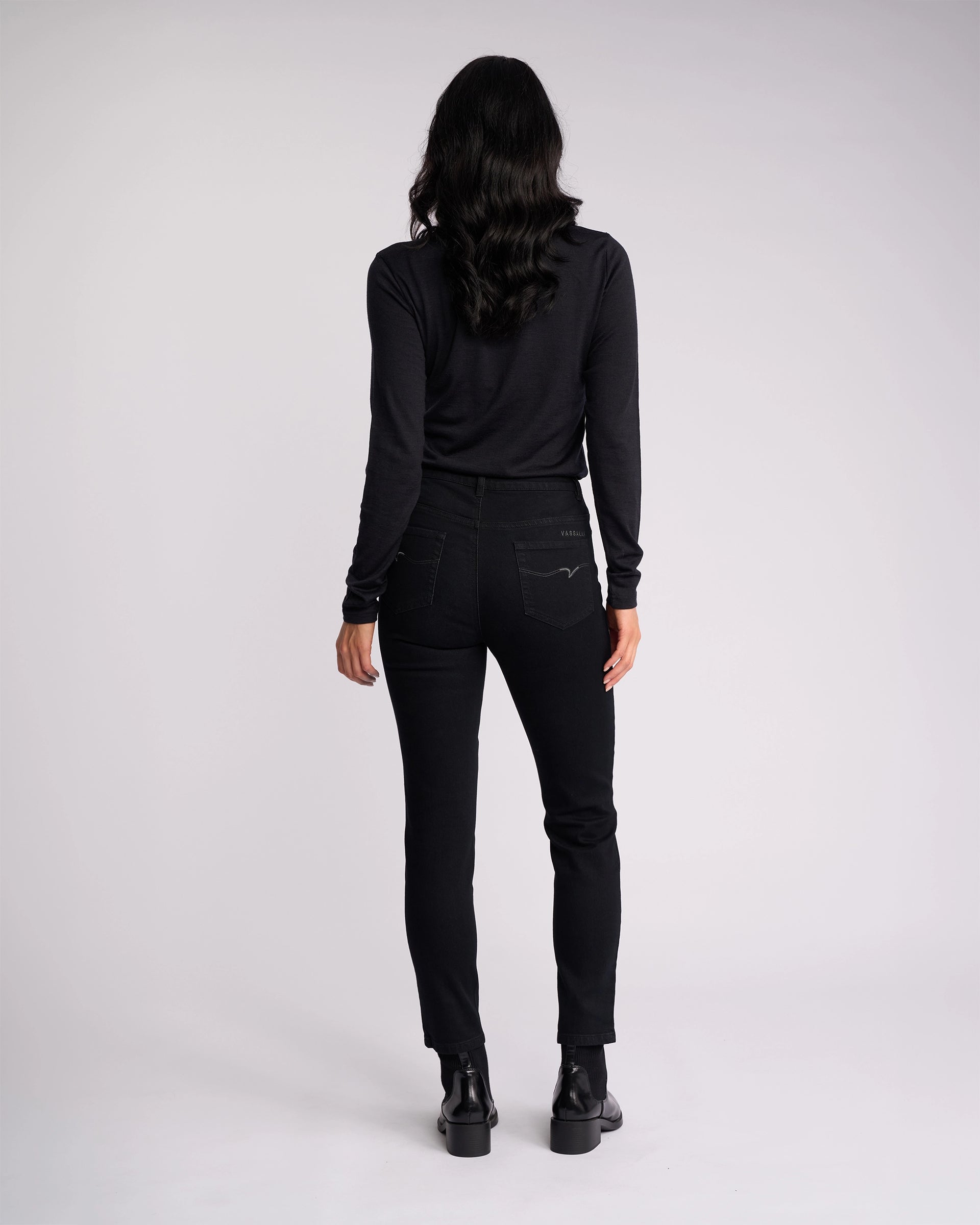 A person with long, wavy hair is seen from behind wearing a black long-sleeve shirt and Vassalli Icon Jean - Black Denim, paired with black shoes, standing against a plain light background.