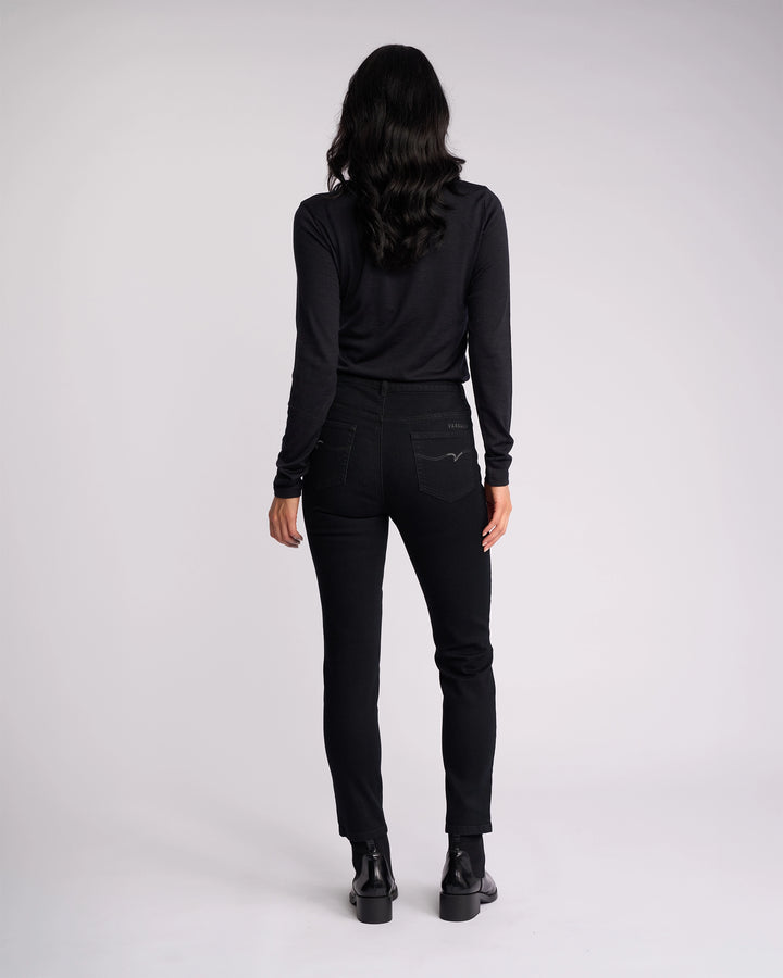 A person with long, wavy hair is seen from behind wearing a black long-sleeve shirt and Vassalli Icon Jean - Black Denim, paired with black shoes, standing against a plain light background.