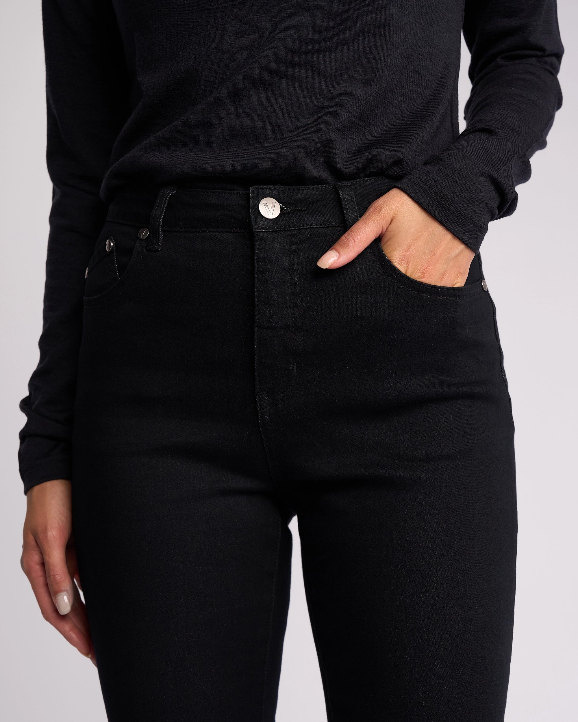 Wearing Vassalli’s Icon Jean in Black Denim, a person stands against a plain background with one hand in their front pocket, styled with a long-sleeved black shirt.