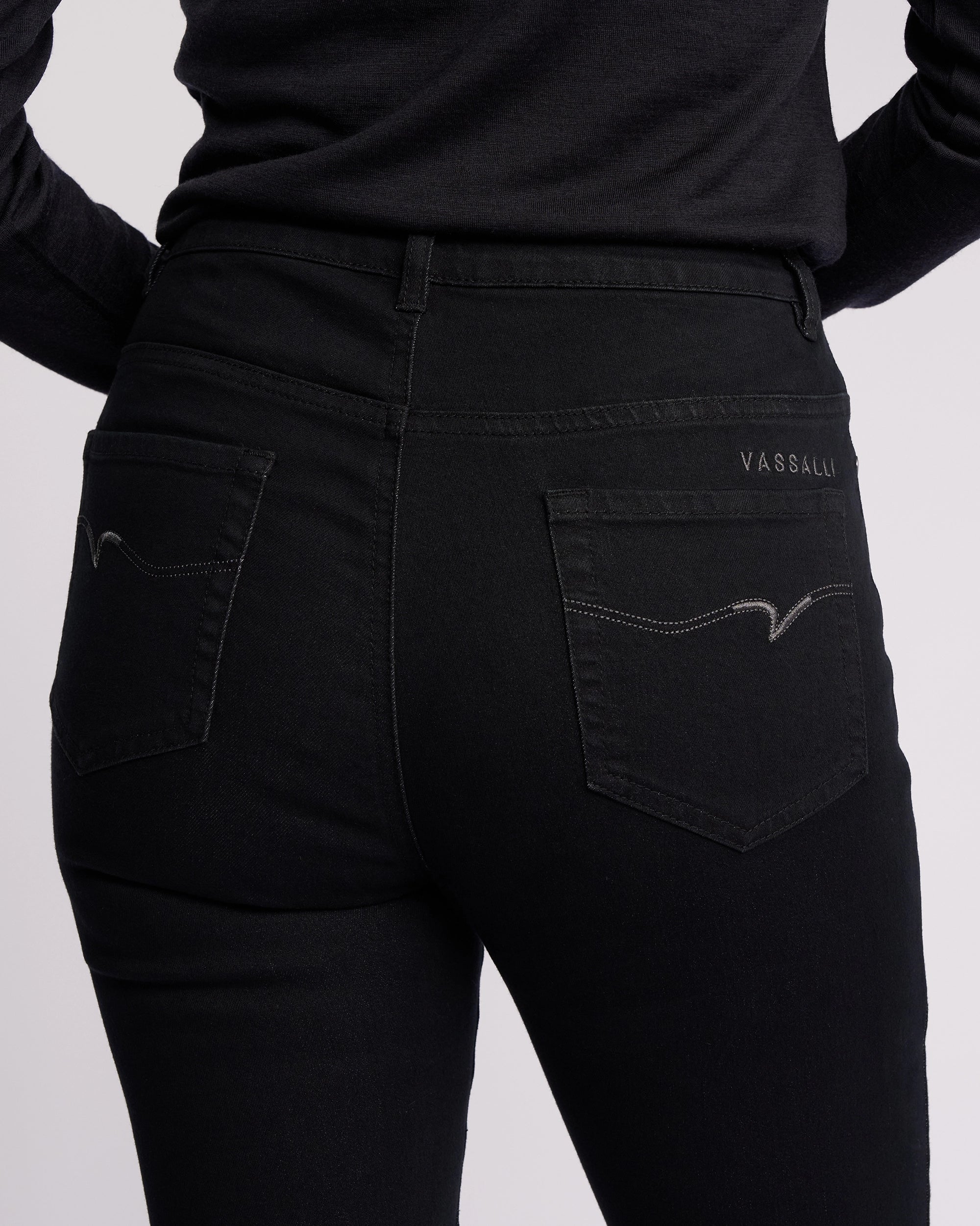 A person wearing Vassalli Icon Jean - Black Denim skinny-leg jeans and a black long-sleeve shirt, shown from the waist down (back view). Jeans feature stretch denim, two back pockets with subtle stitching, and "Vassalli" on the right pocket.