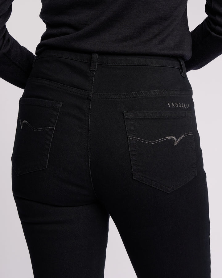 A person wearing Vassalli Icon Jean - Black Denim skinny-leg jeans and a black long-sleeve shirt, shown from the waist down (back view). Jeans feature stretch denim, two back pockets with subtle stitching, and "Vassalli" on the right pocket.