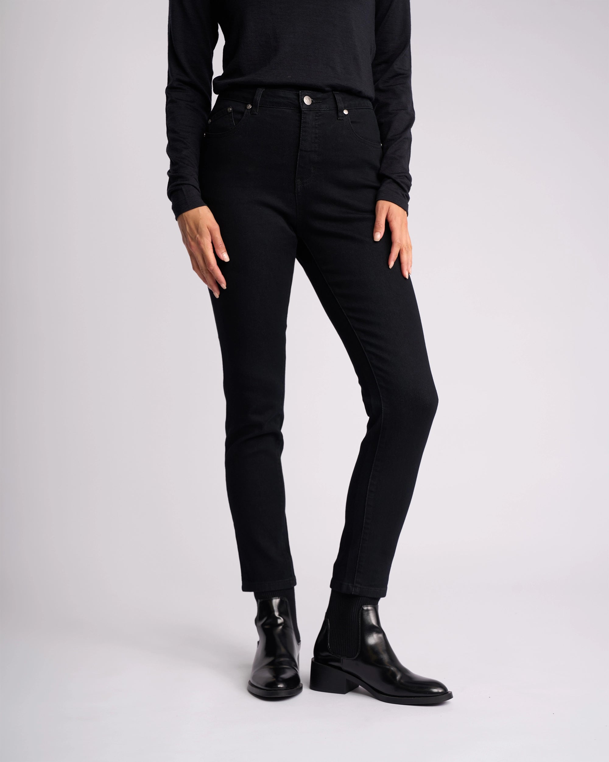 A person models the Vassalli Icon Jean in black denim with a long-sleeve black shirt and black ankle boots against a plain light background. Their hands are at their sides, and their head is out of frame.
