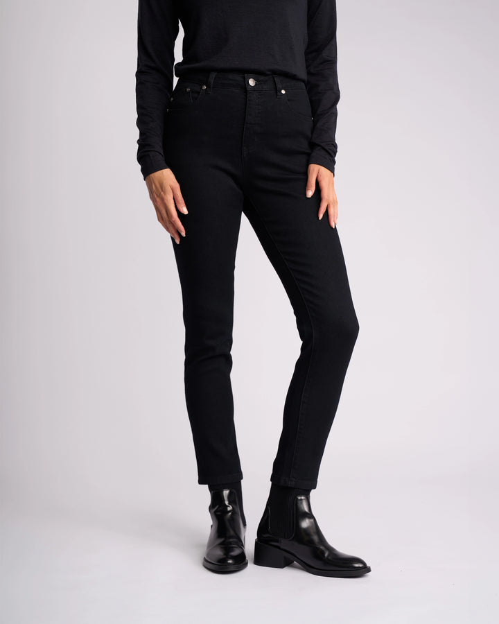 A person models the Vassalli Icon Jean in black denim with a long-sleeve black shirt and black ankle boots against a plain light background. Their hands are at their sides, and their head is out of frame.
