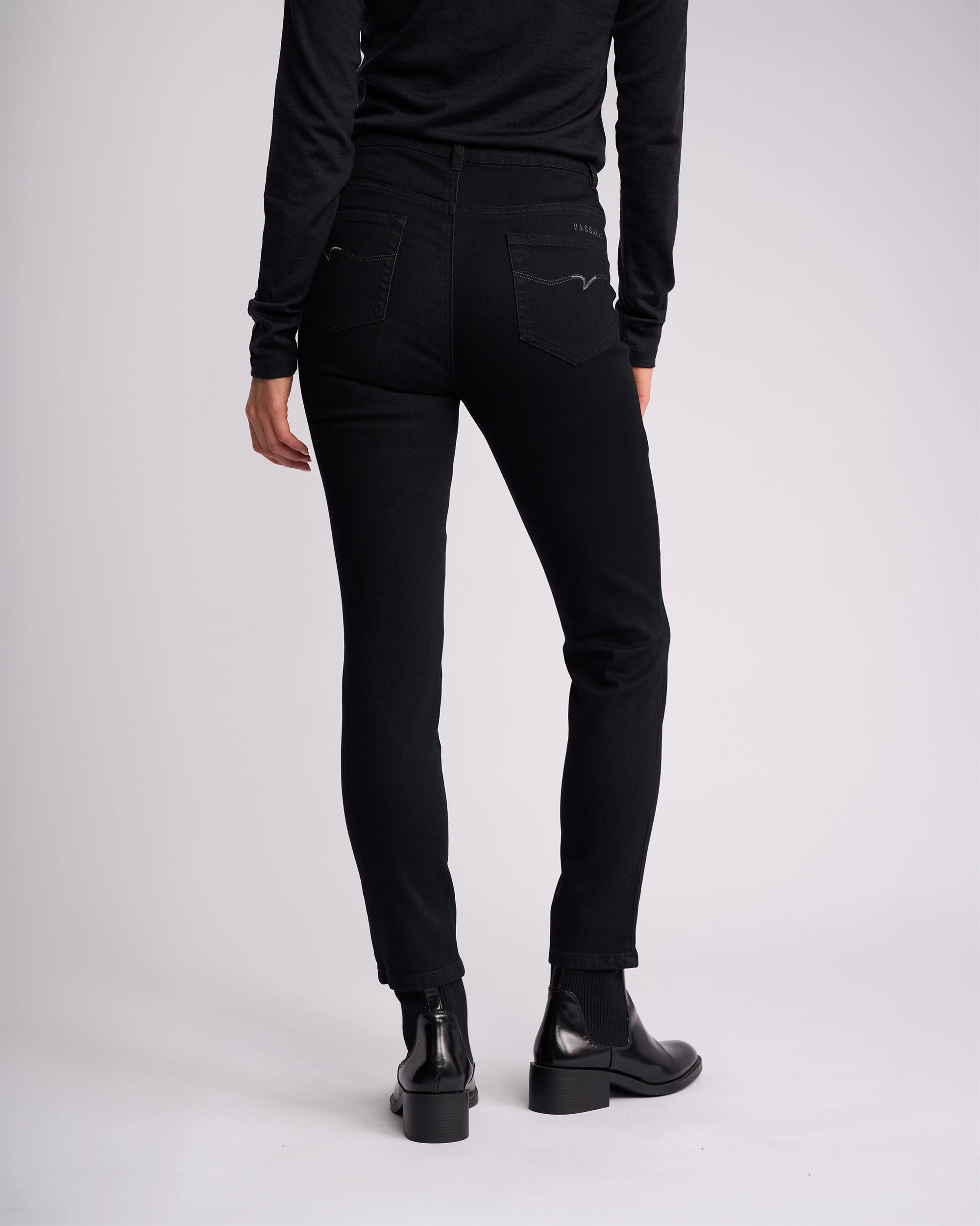 Someone wearing the Vassalli Icon Jean in Black Denim stands with their back to the camera, dressed in a black long-sleeve top and black ankle boots against a plain light background.