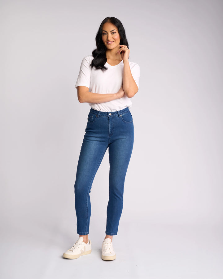 A woman with long dark hair smiles against a plain background, wearing Vassalli's Icon Jean in Blue Denim, a white T-shirt, and white sneakers. She touches her chin with one hand while the other rests by her side.