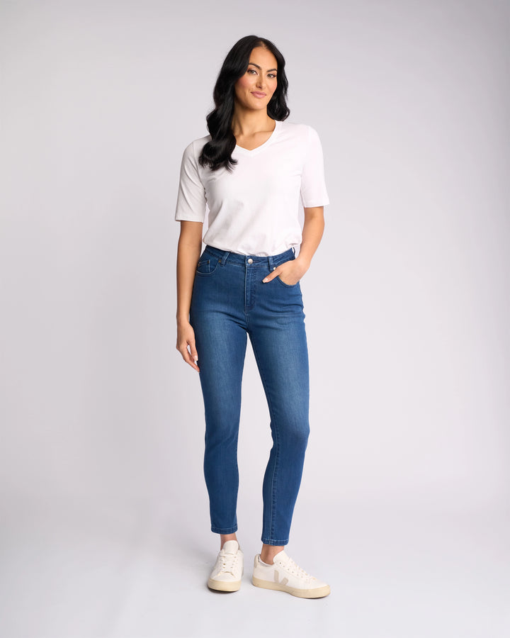 A woman with long dark hair, in a white short-sleeve top and Vassalli Icon Jean – Blue Denim, stands against a plain light background. She wears white sneakers, tucks one hand in her pocket, and smiles slightly.
