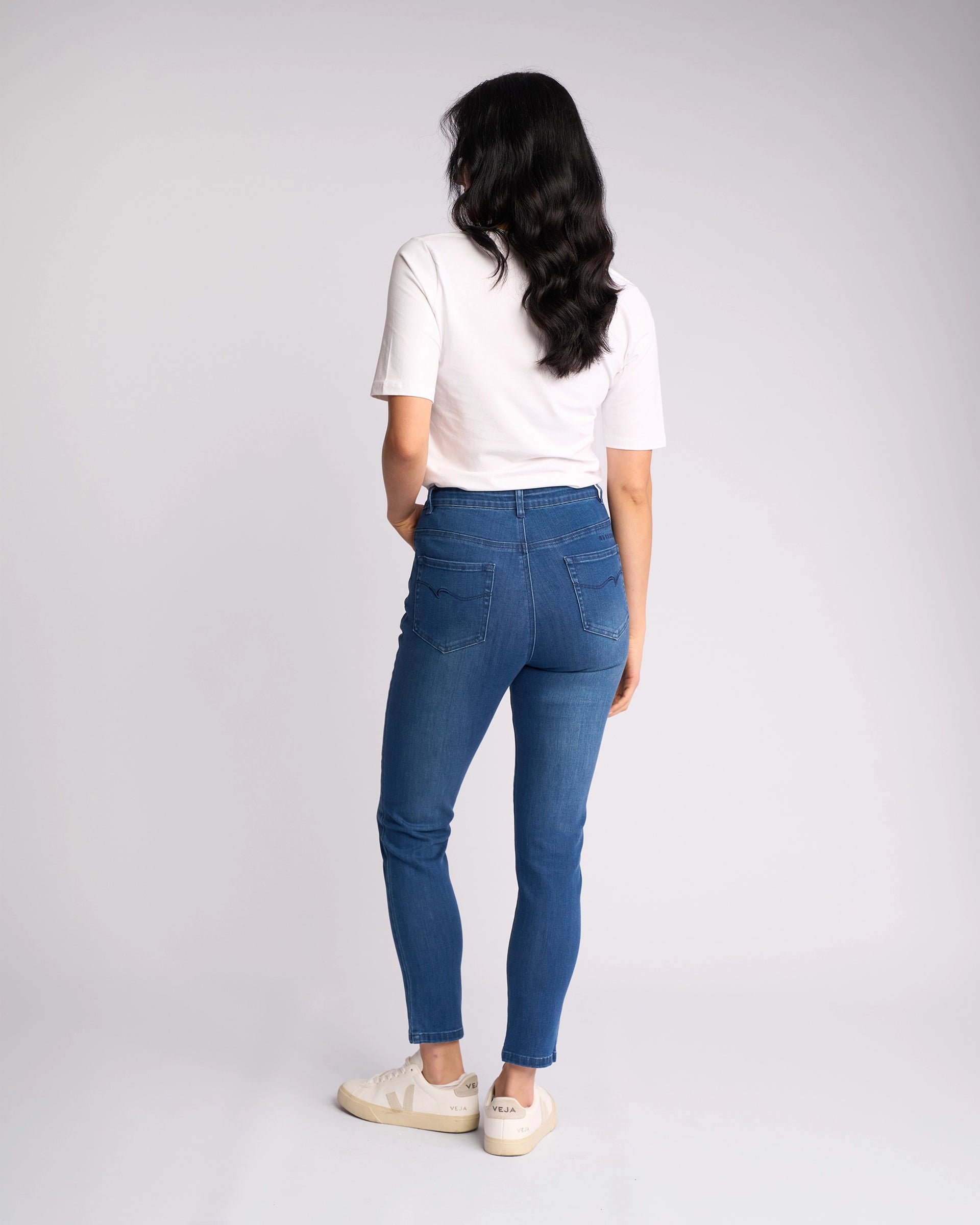 A woman with wavy dark hair poses against a light background, facing away, wearing Vassalli's Icon Jean - Blue Denim, a skinny-leg stretch ankle-grazer, styled with a white short-sleeve shirt and white sneakers.