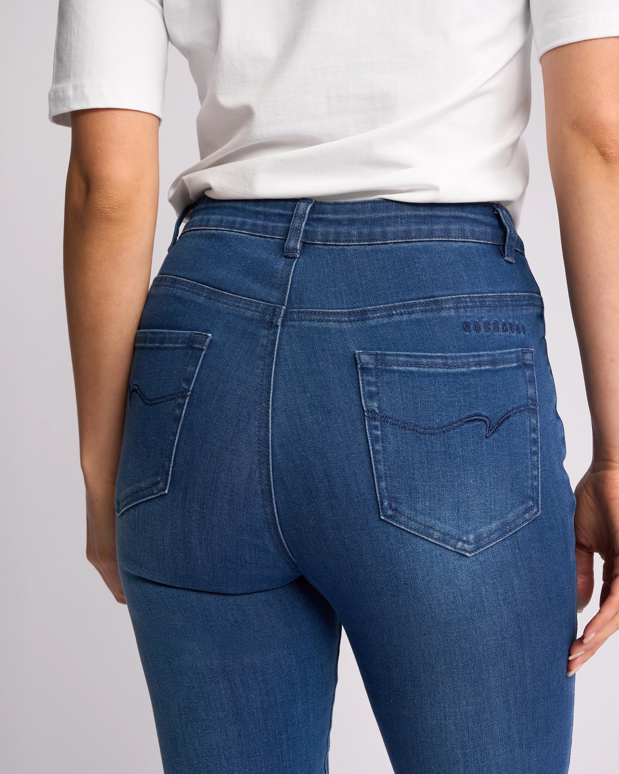 A person is shown from the back wearing Vassalli’s Icon Jean - Blue Denim, highlighting the skinny-leg jeans' back pockets and seam details with a white t-shirt.