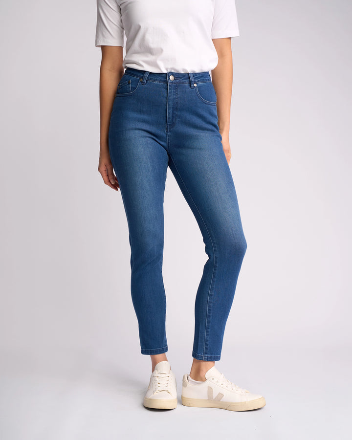 A person in a white t-shirt, Vassalli Icon Jean - Blue Denim ankle-grazer skinny-leg jeans, and white sneakers stands against a plain light background. The person’s face is not visible.