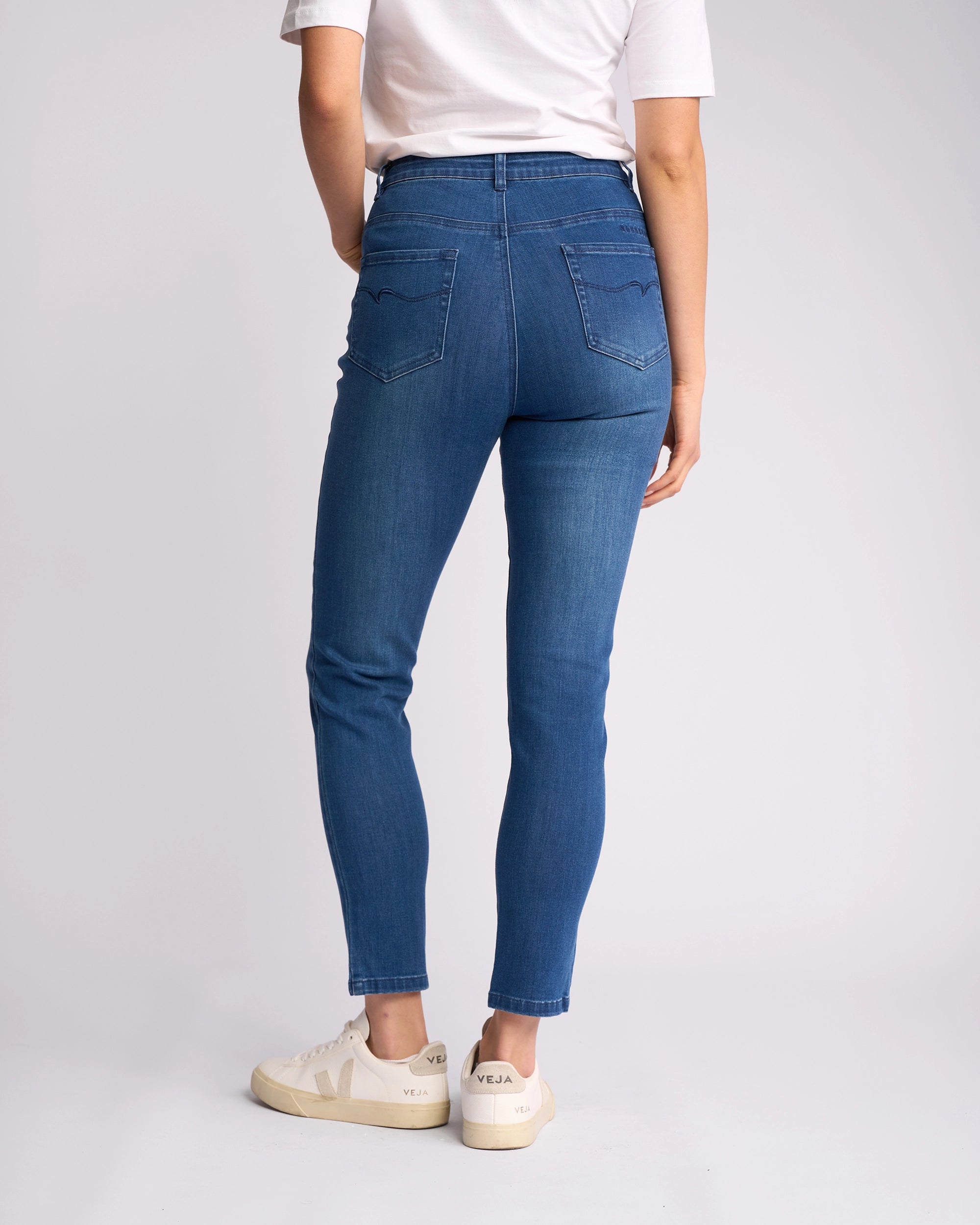 A person faces away, wearing a white t-shirt, Vassalli Icon Jean - Blue Denim high-waisted skinny ankle-grazer jeans in stretch denim, and white Veja sneakers against a plain light background.