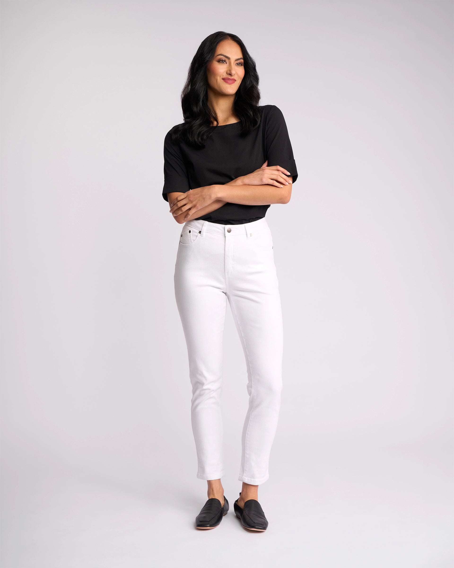 A woman with long dark hair stands against a plain white background, wearing the Vassalli Icon Jean in white denim, a black shirt, and black slip-on shoes. She looks slightly to the side with a relaxed expression.
