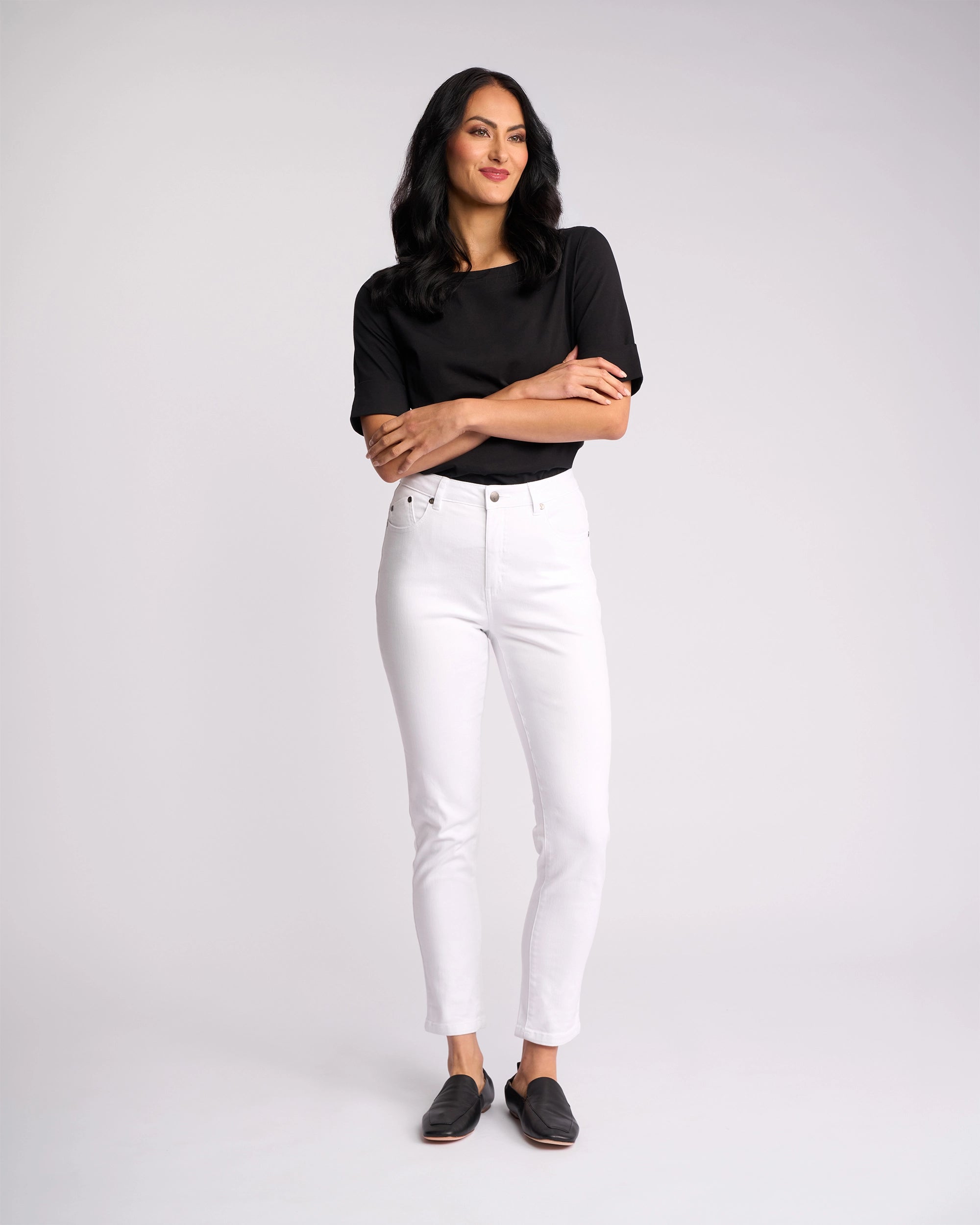A woman with long dark hair stands against a plain white background, wearing the Vassalli Icon Jean in white denim, a black shirt, and black slip-on shoes. She looks slightly to the side with a relaxed expression.