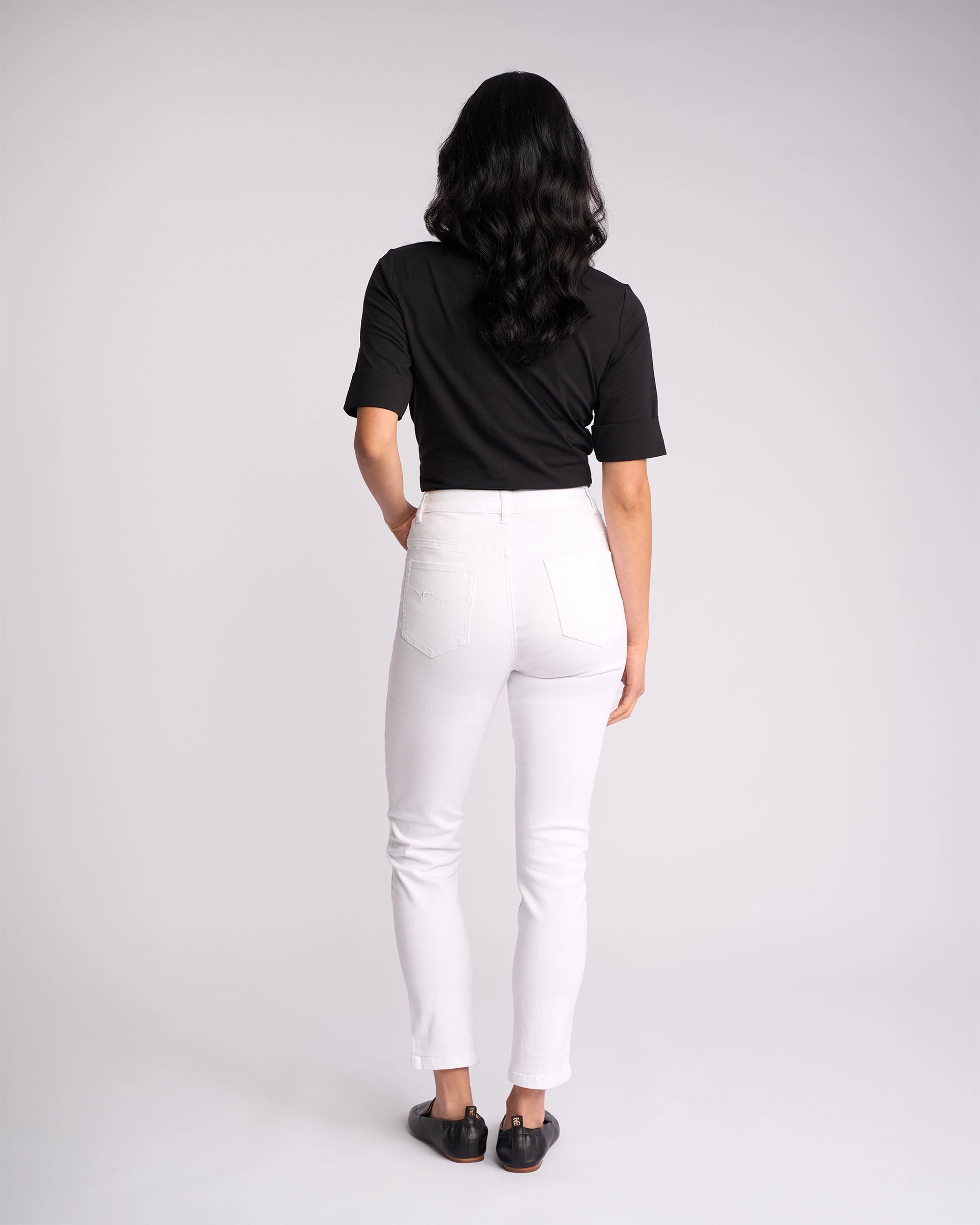 A person with long, dark hair stands facing away against a plain light background, wearing a black short-sleeve shirt, black shoes, and Vassalli’s Icon Jean - White Denim.