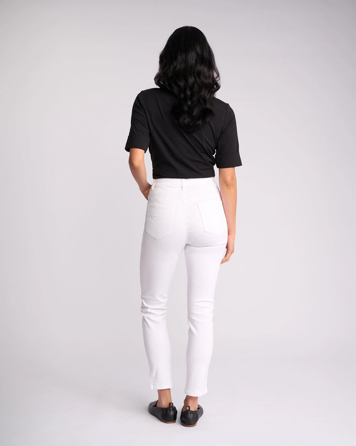 A person with long, dark hair stands facing away against a plain light background, wearing a black short-sleeve shirt, black shoes, and Vassalli’s Icon Jean - White Denim.