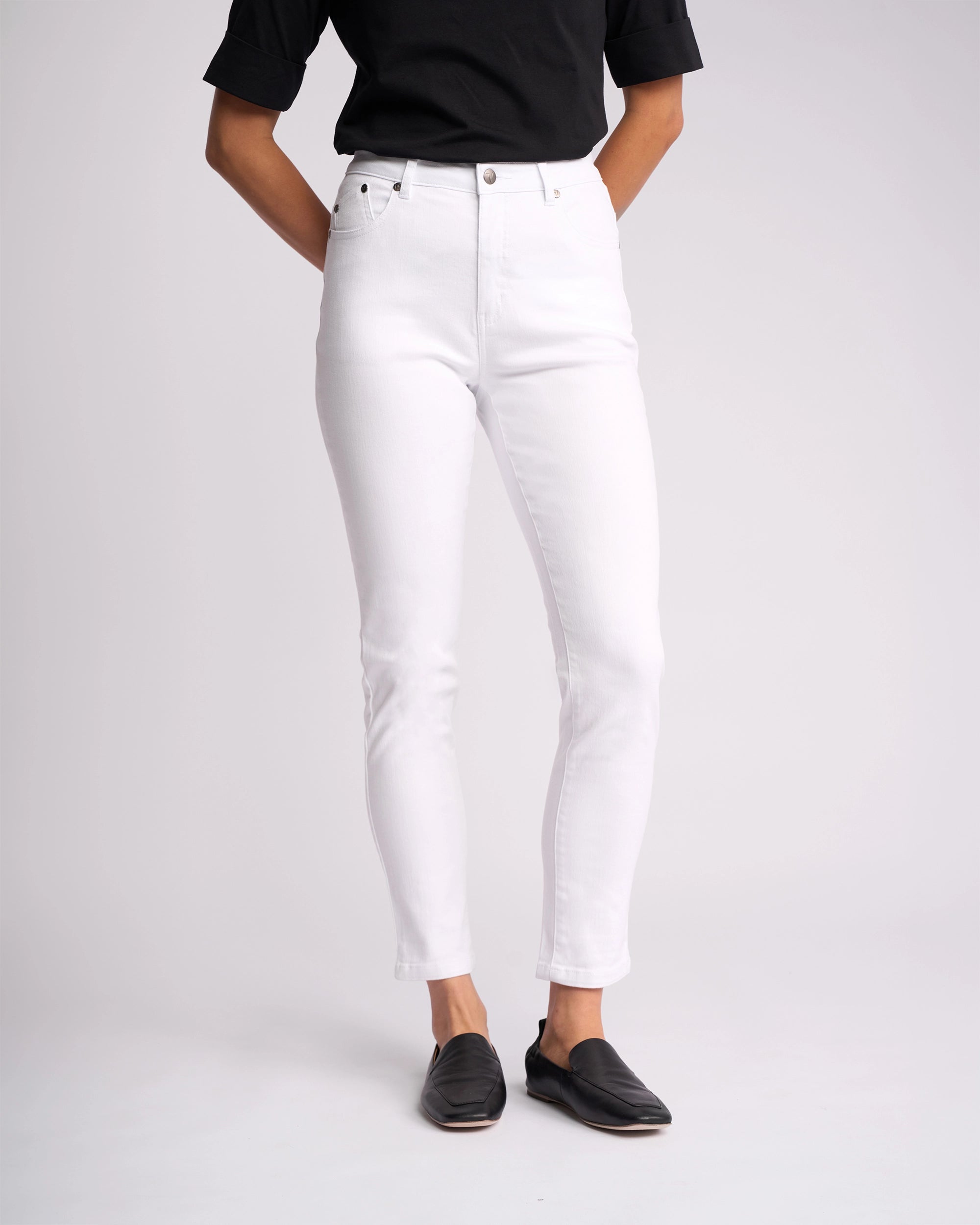 Person wearing a black short-sleeve top, Vassalli Icon Jean - White Denim (high-waisted, skinny-leg), and black slip-on shoes, standing against a plain light background. The person's face is not visible.
