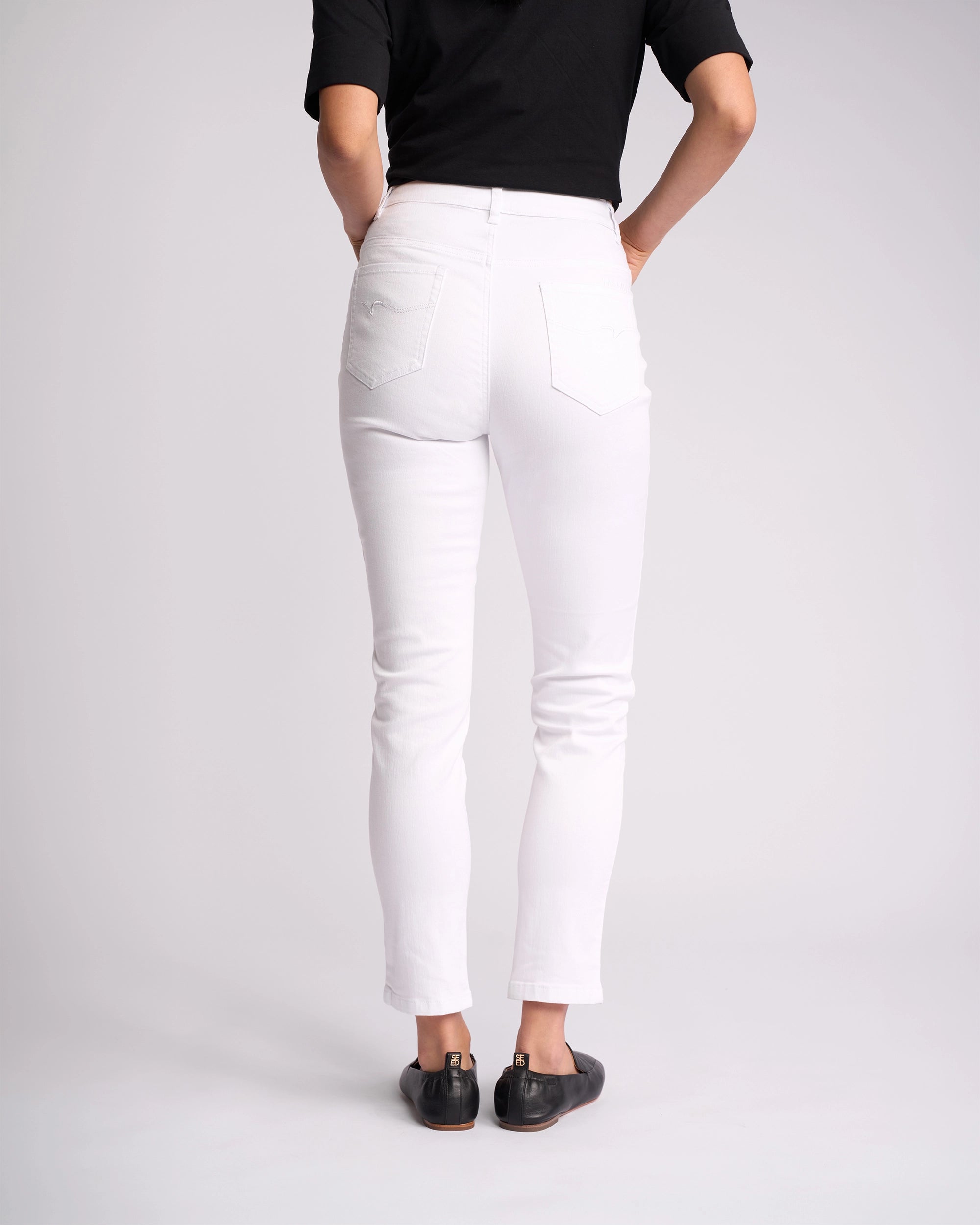 A person stands facing away, wearing a black short-sleeve top, black flats, and Vassalli Icon Jean in White Denim against a plain light background.