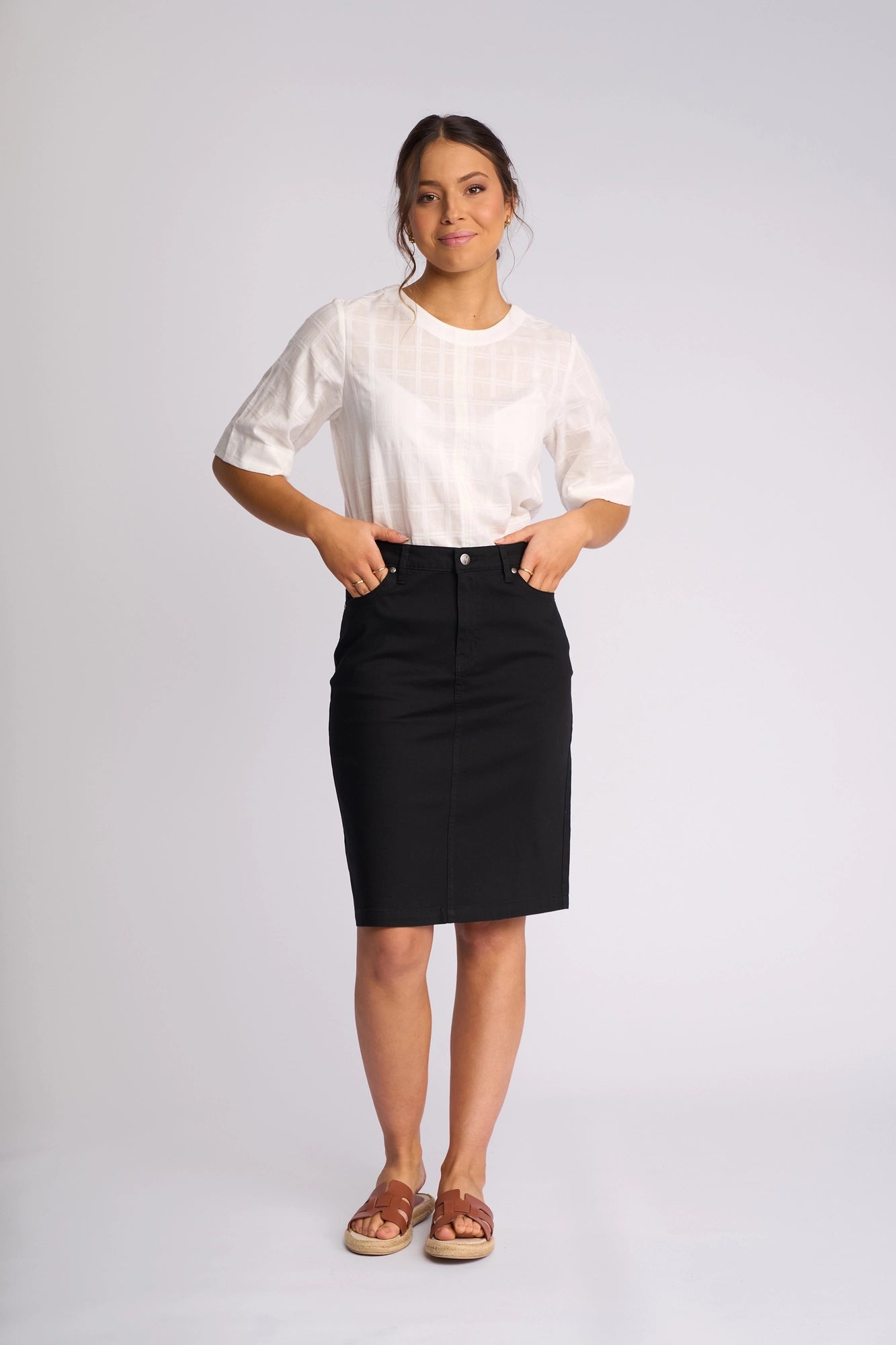A woman stands against a plain background, wearing a white short-sleeve blouse paired with the Vassalli Tilt Skirt in black, crafted from stretch cotton. She completes her look with brown sandals, placing her hands in the pockets of her skirt and smiling slightly at the camera.