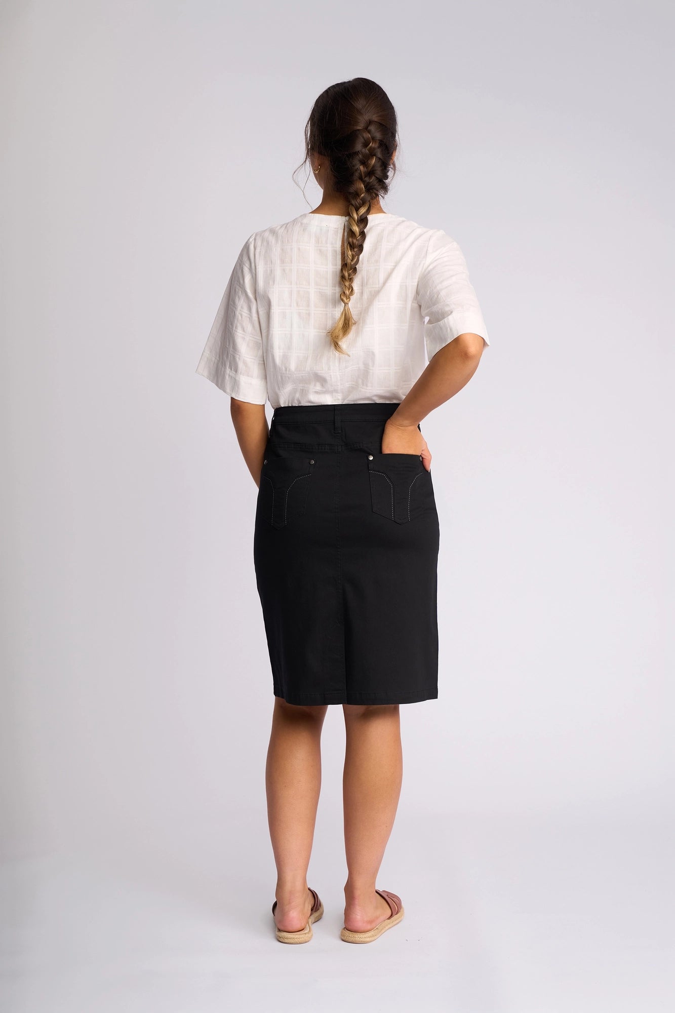 A woman with a braid stands facing away, dressed in a white short-sleeve blouse and the Vassalli Tilt Skirt – Black. The knee-length skirt, made from stretch cotton for comfort, features practical pockets. She completes her look with sandals and slips one hand into her pocket, posed against a simple light background.