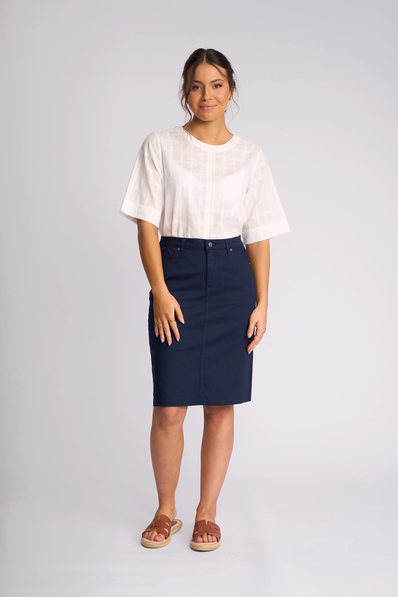 A woman stands against a plain background, dressed in a white short-sleeve blouse and the Vassalli Tilt Skirt in navy—a comfortable piece crafted from soft stretch cotton. She completes her look with brown sandals and offers a gentle smile, her arms resting naturally at her sides.