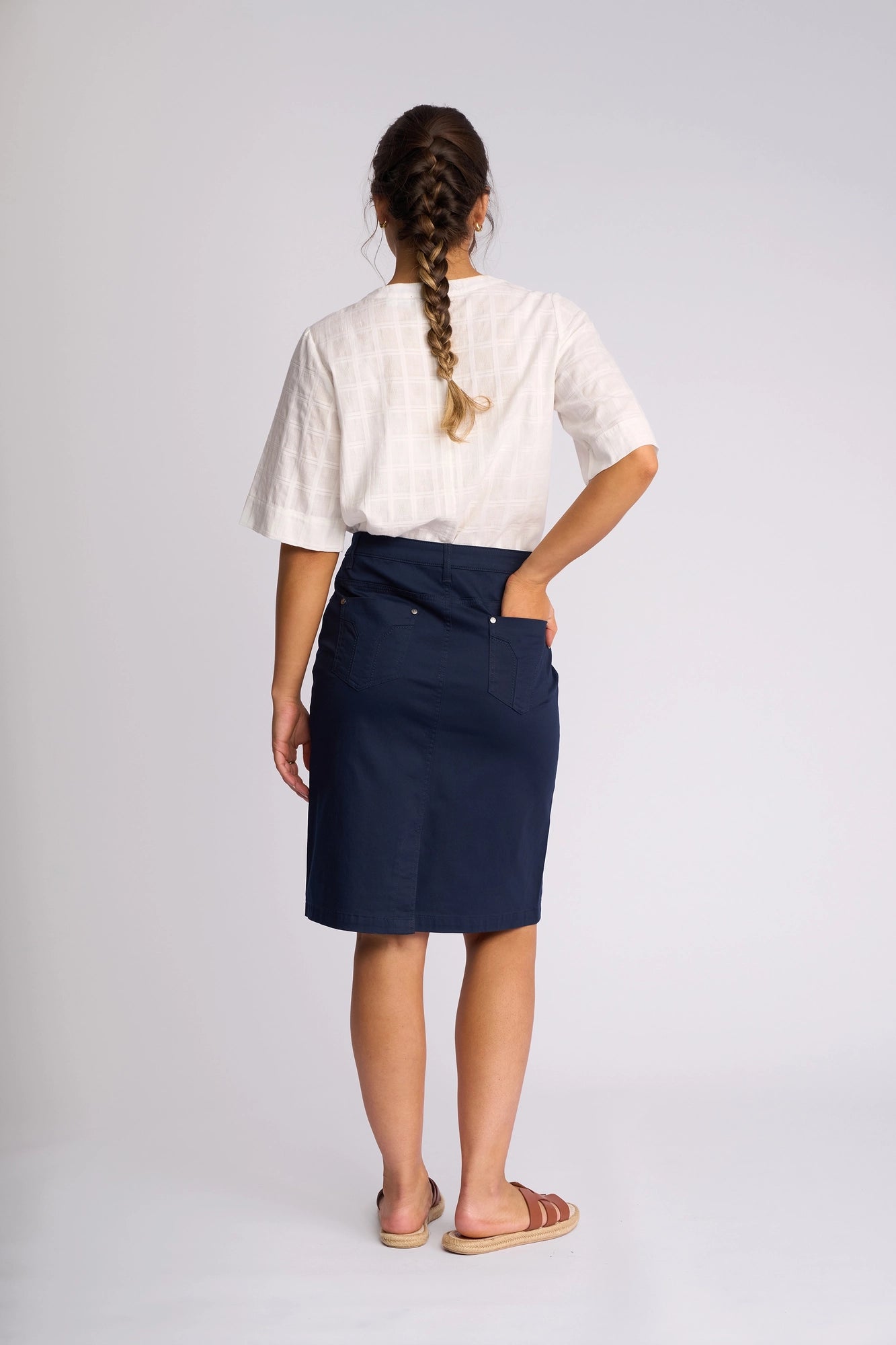 A woman with a long braid, dressed in a white short-sleeve top, the Vassalli Tilt Skirt - Navy that falls to her knees, and sandals, stands with her back to the camera and one hand in her pocket against a plain light background.