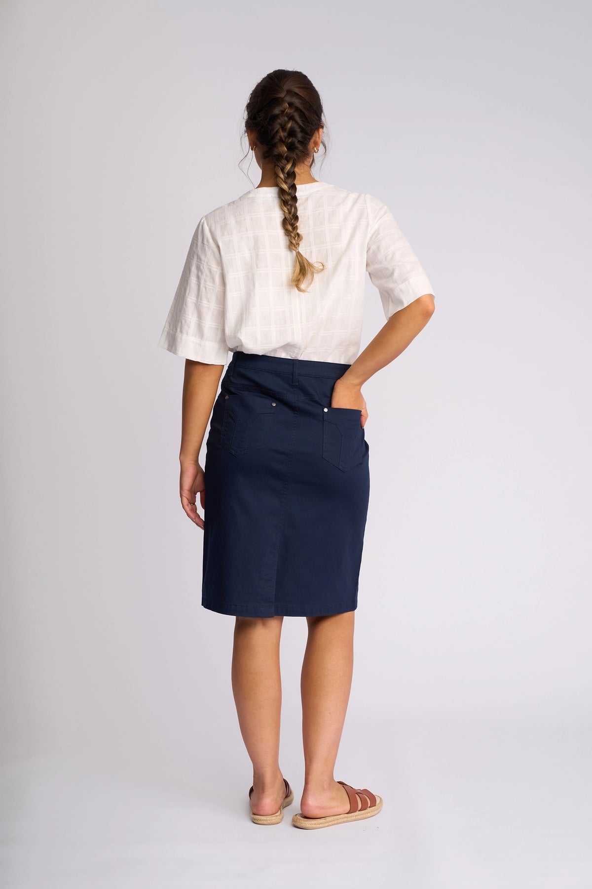 A woman with a long braid, dressed in a white short-sleeve top, the Vassalli Tilt Skirt - Navy that falls to her knees, and sandals, stands with her back to the camera and one hand in her pocket against a plain light background.