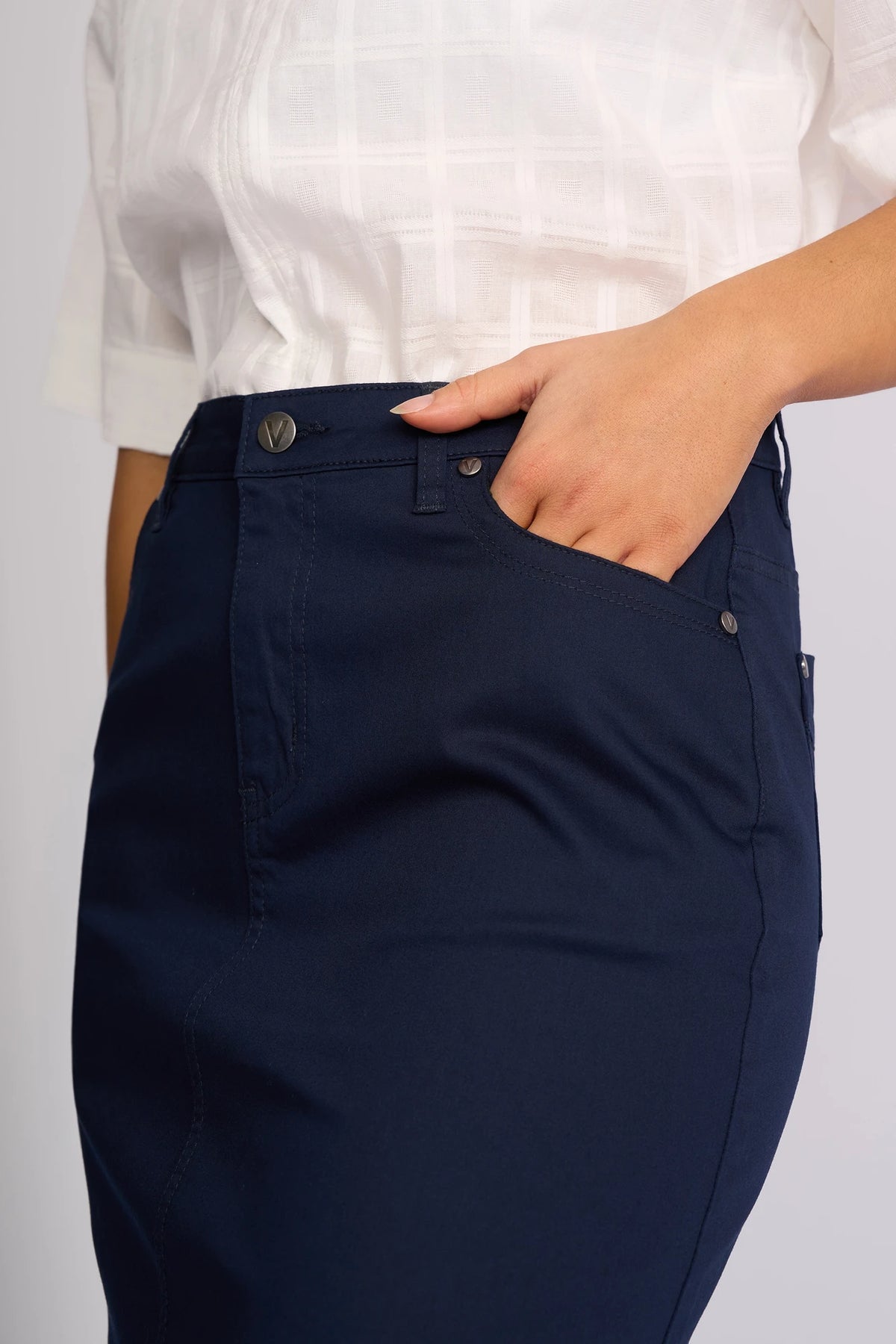 A person wearing a white short-sleeve blouse and a navy skirt, such as the Vassalli Tilt Skirt in Navy, stands with one hand resting in their front pocket.
