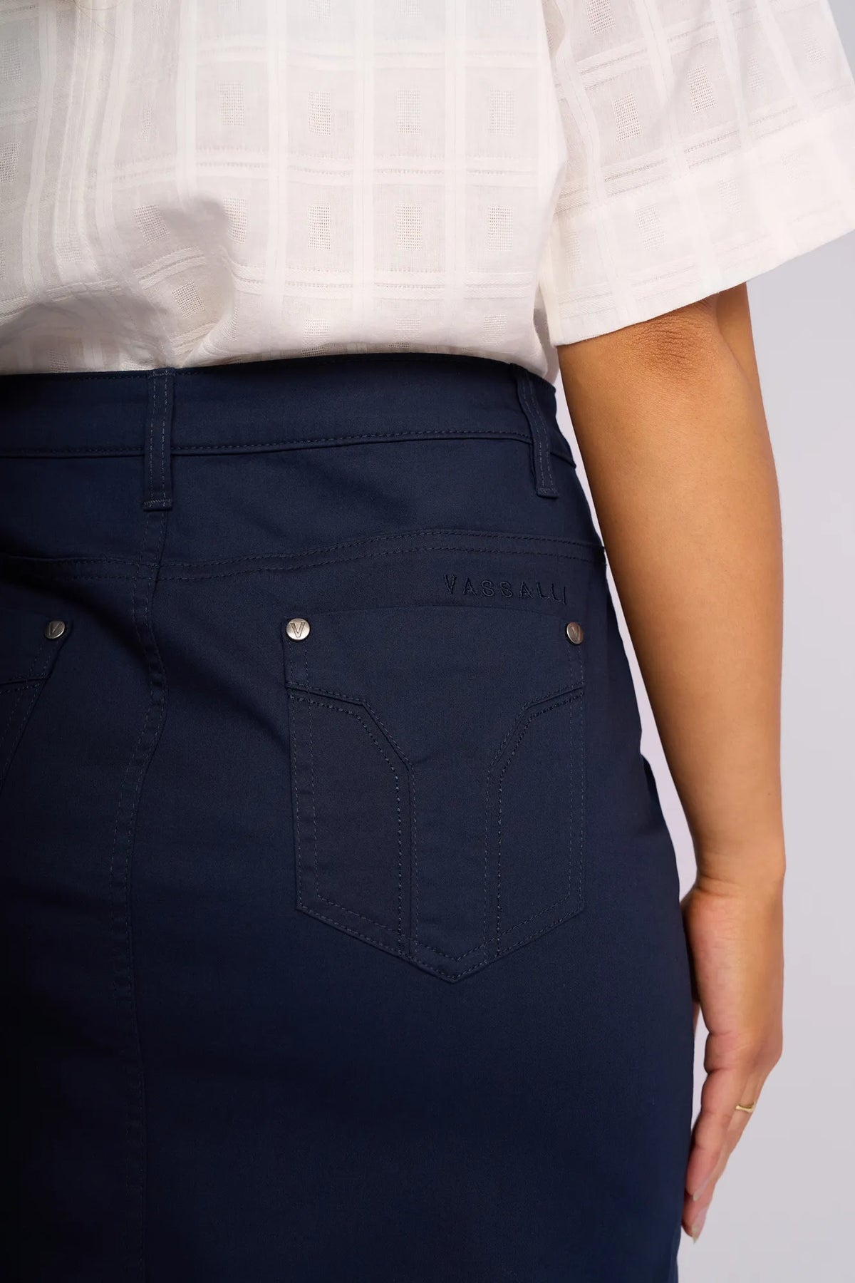 A person is shown from the waist down wearing the Vassalli Tilt Skirt in navy, paired with a white short-sleeve blouse, with their right hand resting by their side.