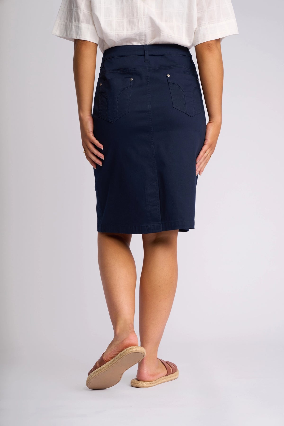 A person stands facing away, wearing a white short-sleeve shirt, the Vassalli Tilt Skirt in navy—a knee-length stretch cotton skirt featuring back pockets—and flat sandals against a plain background.
