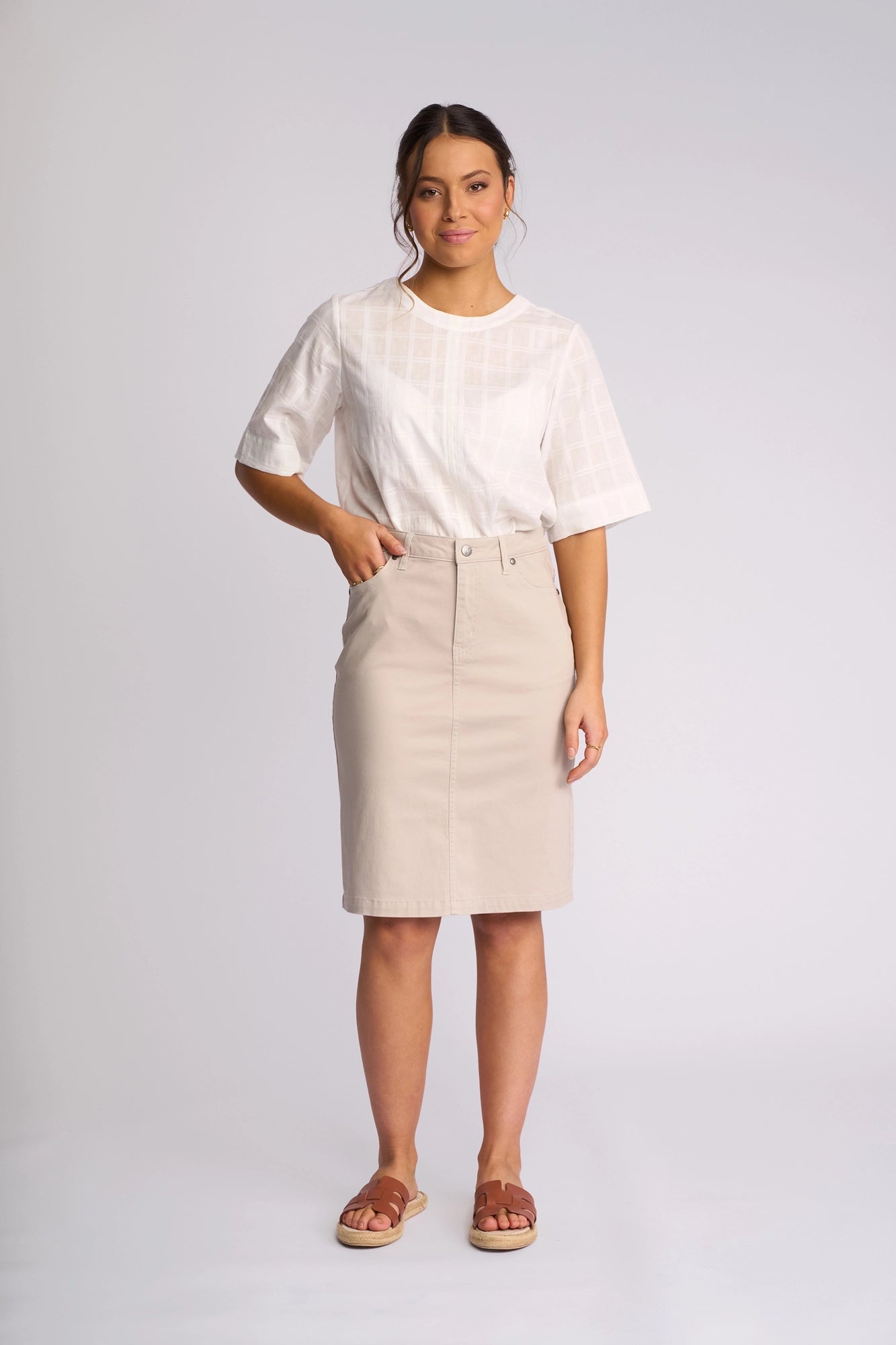 A woman stands against a plain light background, wearing a white short-sleeve blouse, the Vassalli Tilt Skirt in Pumice, and brown sandals. Her hair is tied back and she has a soft, neutral expression.