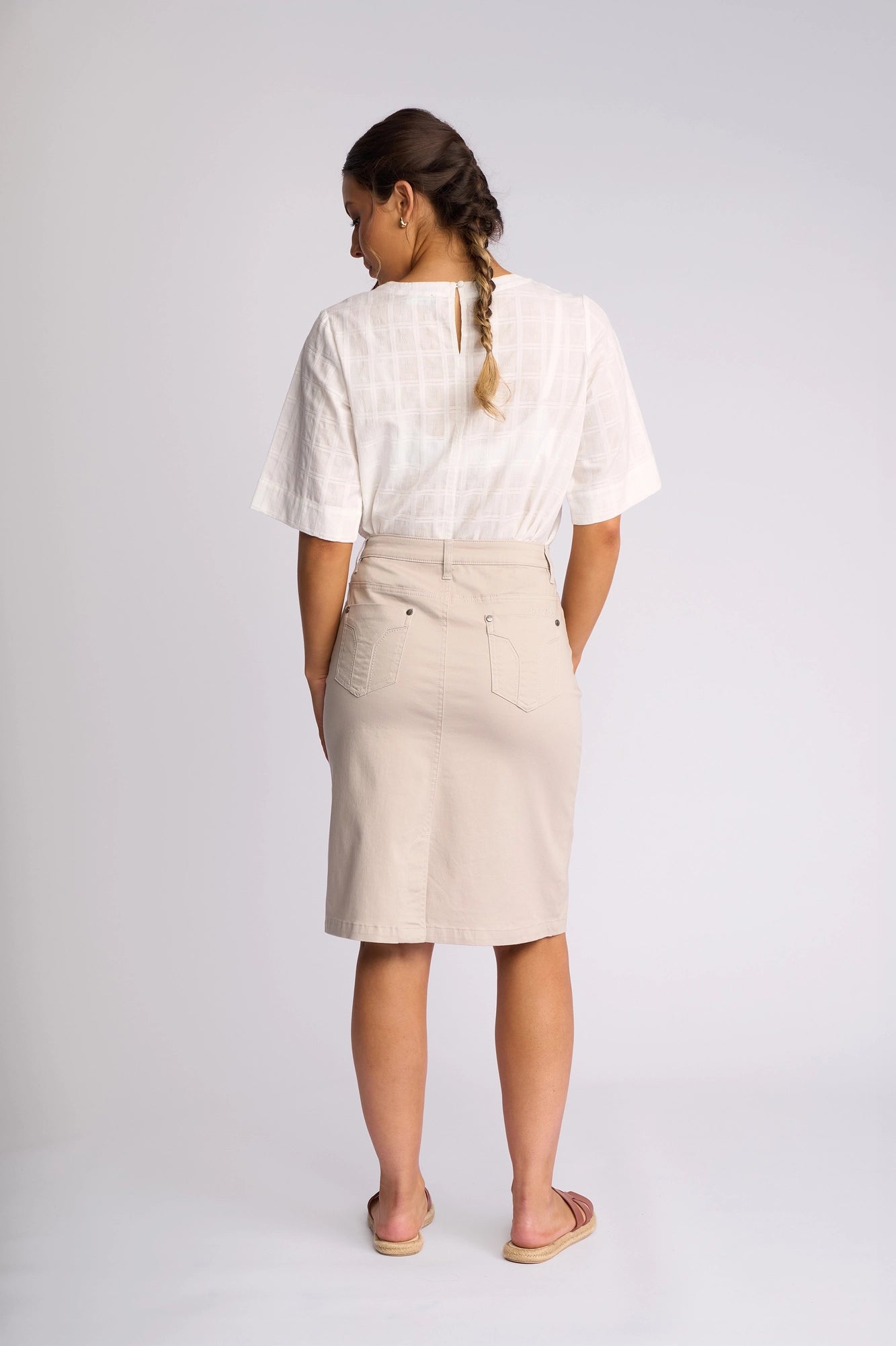 A woman stands with her back to the camera, wearing a white short-sleeve blouse, sandals, and the Vassalli Tilt Skirt in Pumice, crafted from stretch cotton for comfort. Her hair is braided, and the background is plain white.