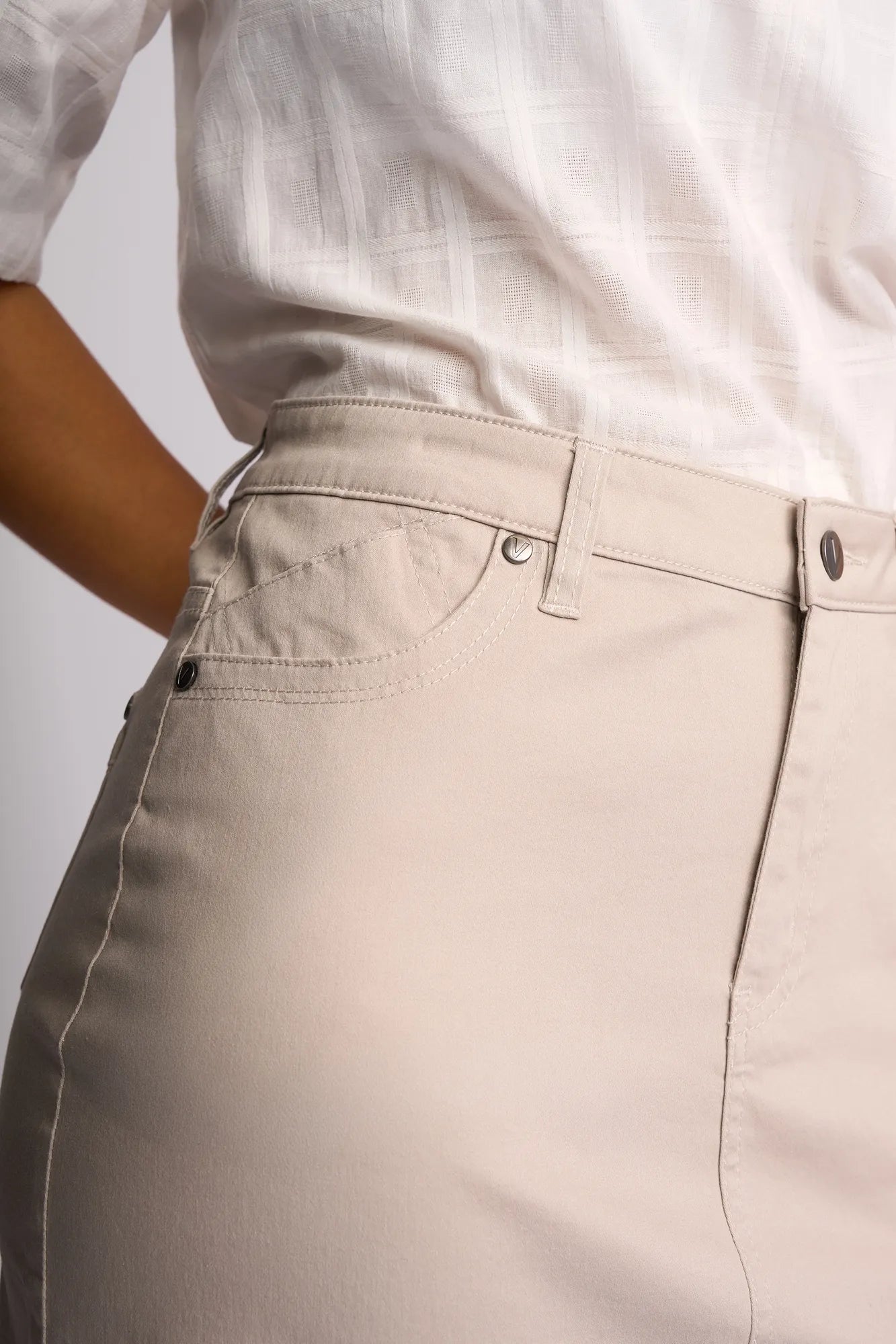 A person wearing a white textured blouse and a high-waisted Vassalli Tilt Skirt in Pumice, crafted from beige stretch cotton, with one hand resting on their hip, shown from the mid-torso to upper thigh.