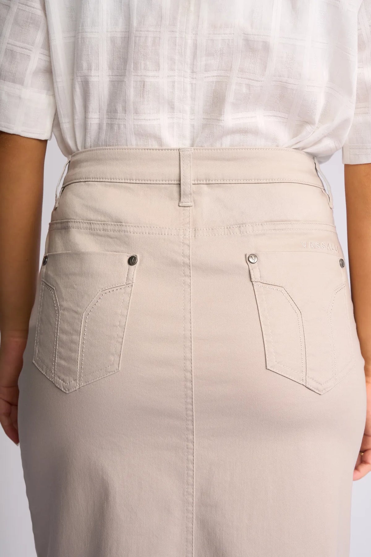 Person wearing a white textured short-sleeve shirt tucked into a beige Vassalli Tilt Skirt in Pumice, featuring two back pockets and belt loops, viewed from behind.