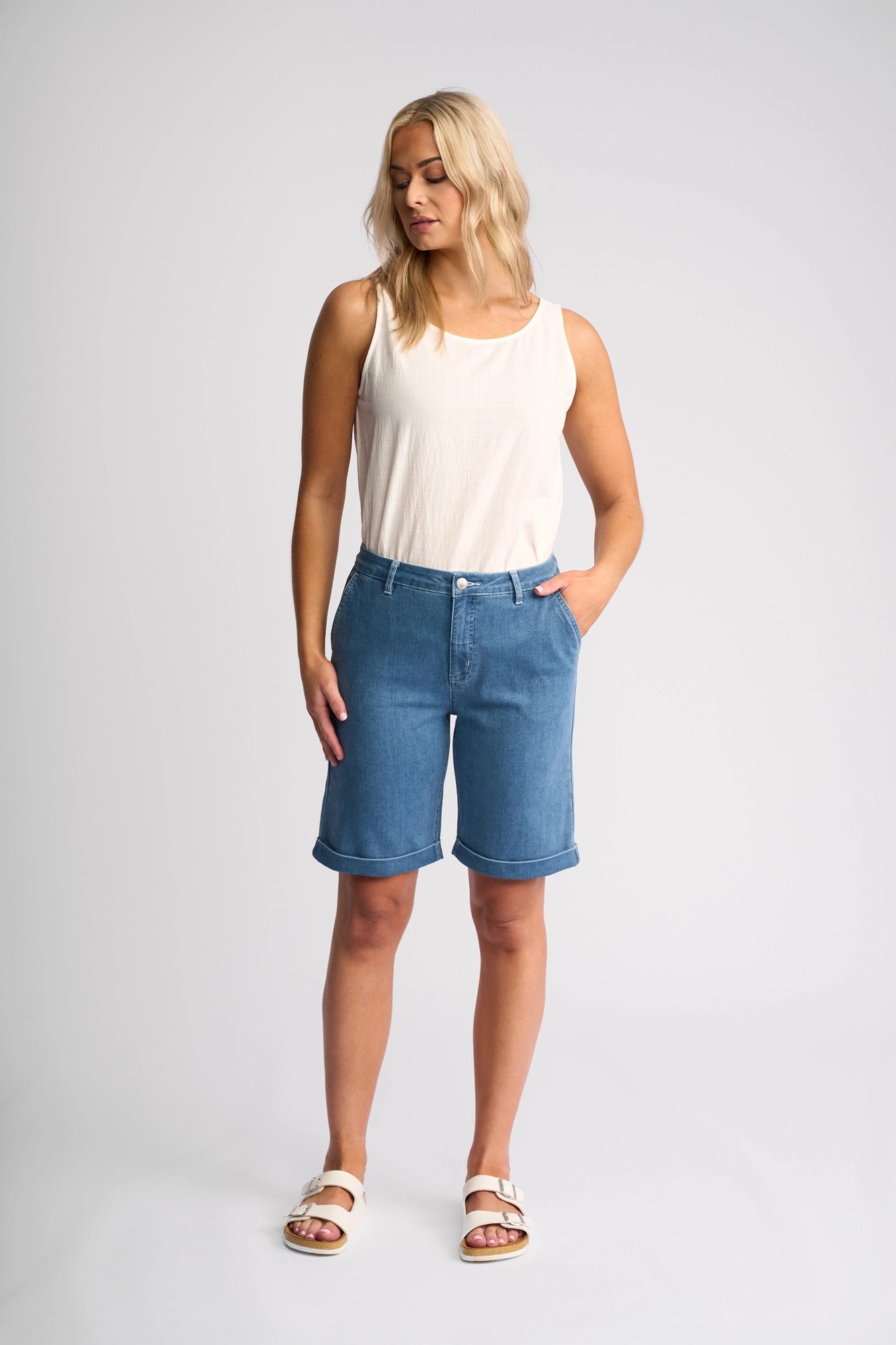 A woman wearing Vassalli’s Havana Short in Blue Hue Denim—a true summer staple—pairs the knee-length stretch shorts with a sleeveless white top and white sandals. She stands against a plain light background, one hand in her pocket, looking down to the side.