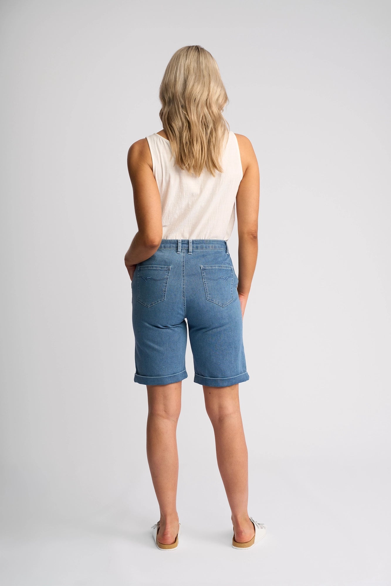 A person with blonde hair, dressed in a white sleeveless top and the Vassalli Havana Short in Blue Hue Denim—a true summer staple—stands facing away from the camera against a plain light background. Sandals complete the relaxed look.