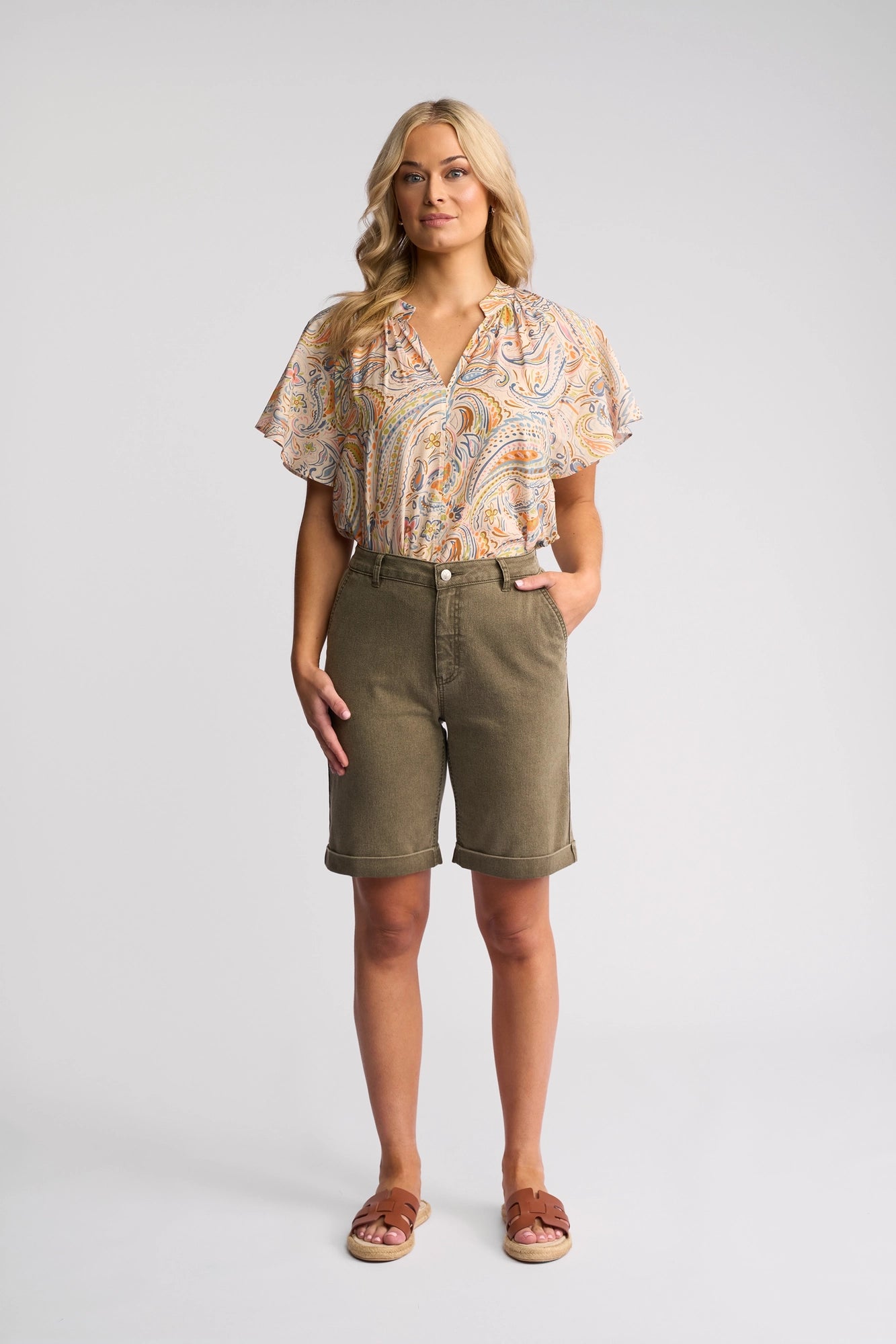A woman stands against a neutral background wearing the Vassalli Havana Short in Sage Stripe Denim—a perfect summer staple—paired with a paisley short-sleeve blouse and brown sandals. She has long, wavy blonde hair and one hand in her pocket.