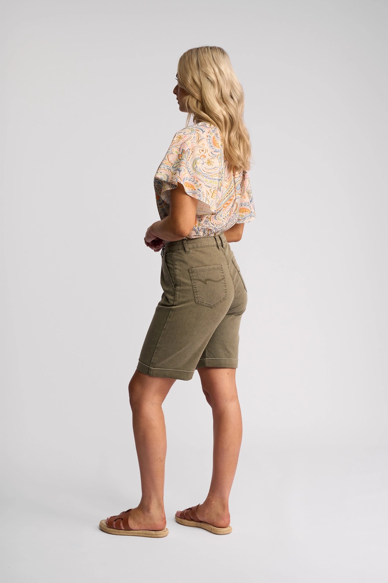A woman with long blonde hair poses sideways against a plain white background, wearing the Vassalli Havana Short in Sage Stripe Denim—her go-to summer staple—paired with an orange patterned top and sandals.
