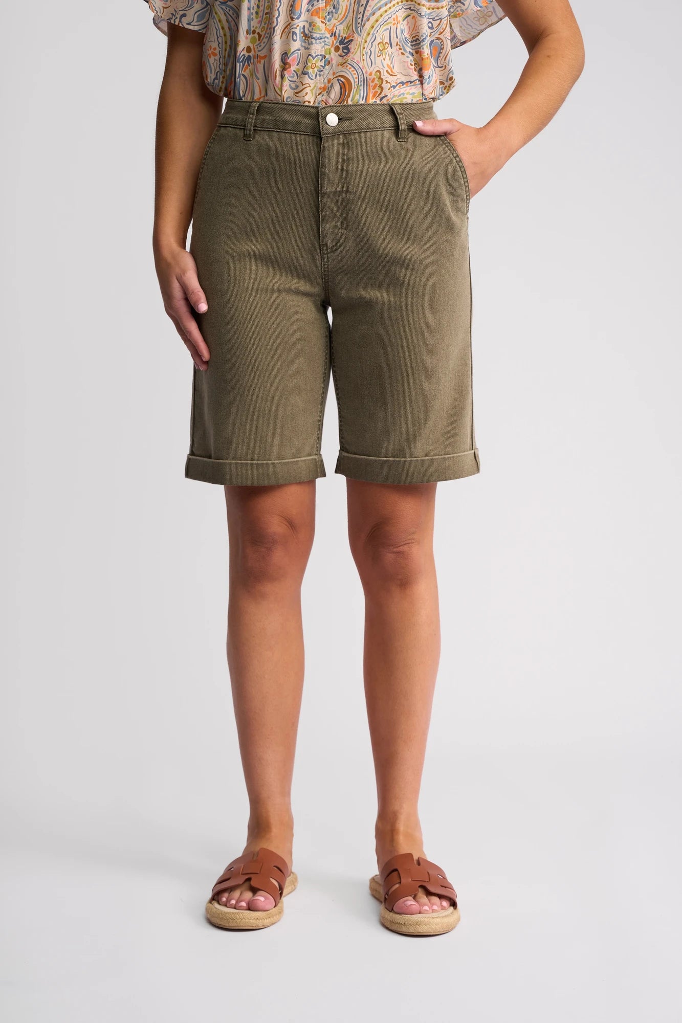 A person models the Vassalli Havana Short in Sage Stripe Denim—olive green, knee-length shorts—paired with a paisley patterned top and brown sandals, standing against a plain light background with hands in their pockets: a fresh take on summer style that goes beyond the classic denim short.
