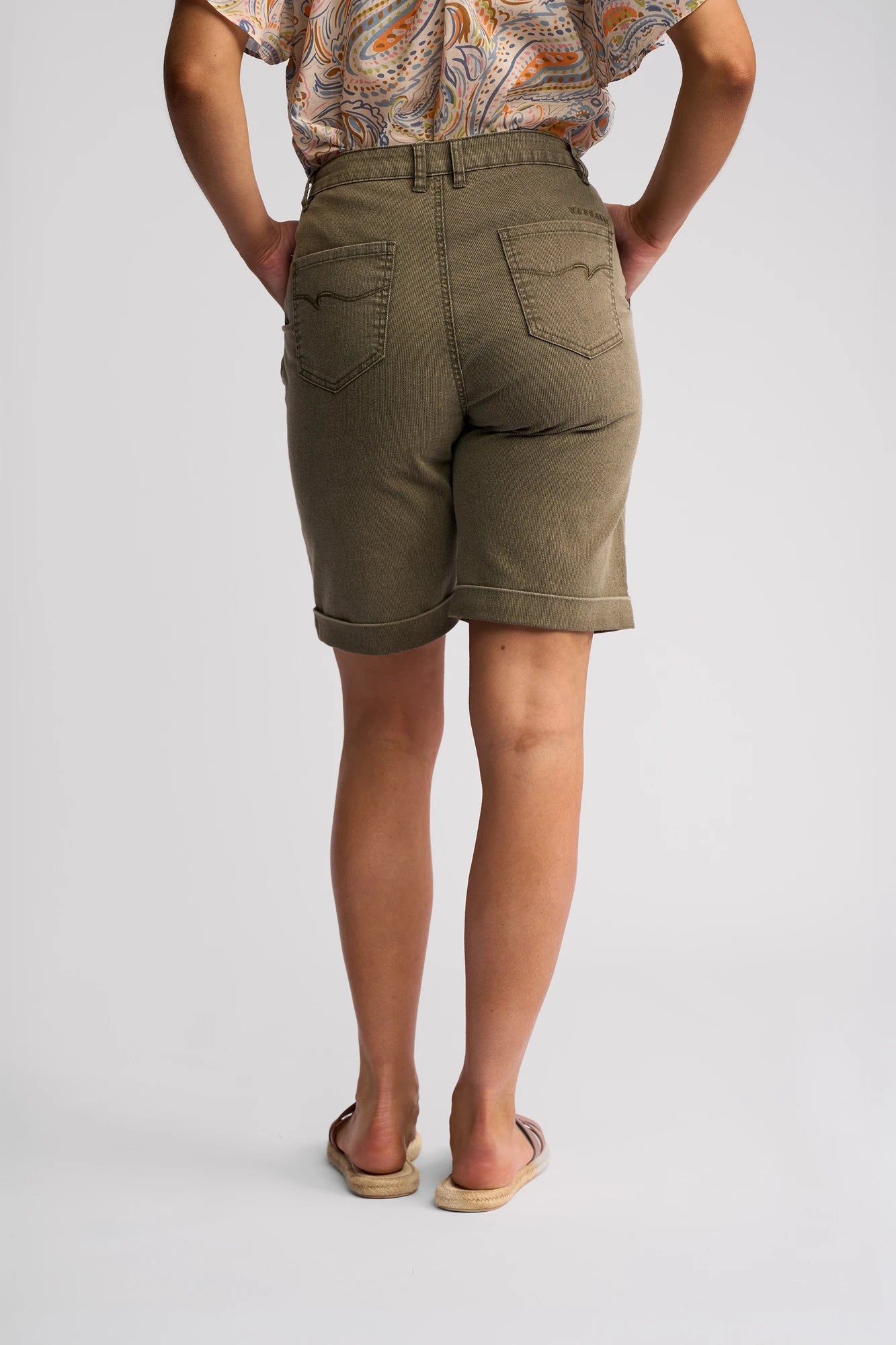 A person wearing the Vassalli Havana Short in Sage Stripe Denim, paired with a patterned top and tan slip-on shoes, stands facing away with their hands in their back pockets against a plain light background—a true summer staple look.