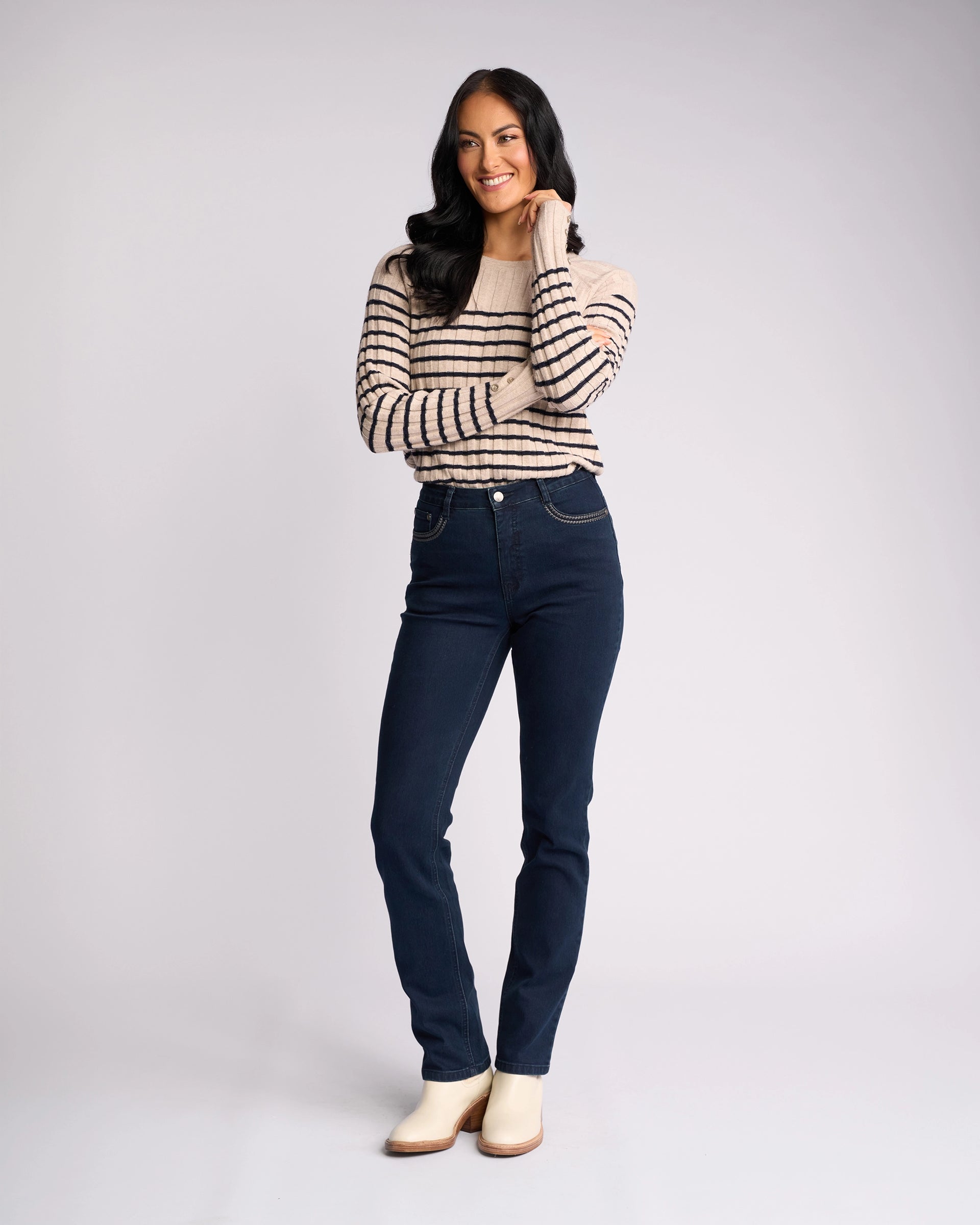 A woman with long dark hair smiles, touching her face, wearing a beige striped sweater, Vassalli’s Urban Jean - Mid Blue high-rise slim-leg jeans, and light ankle boots against a plain light background.