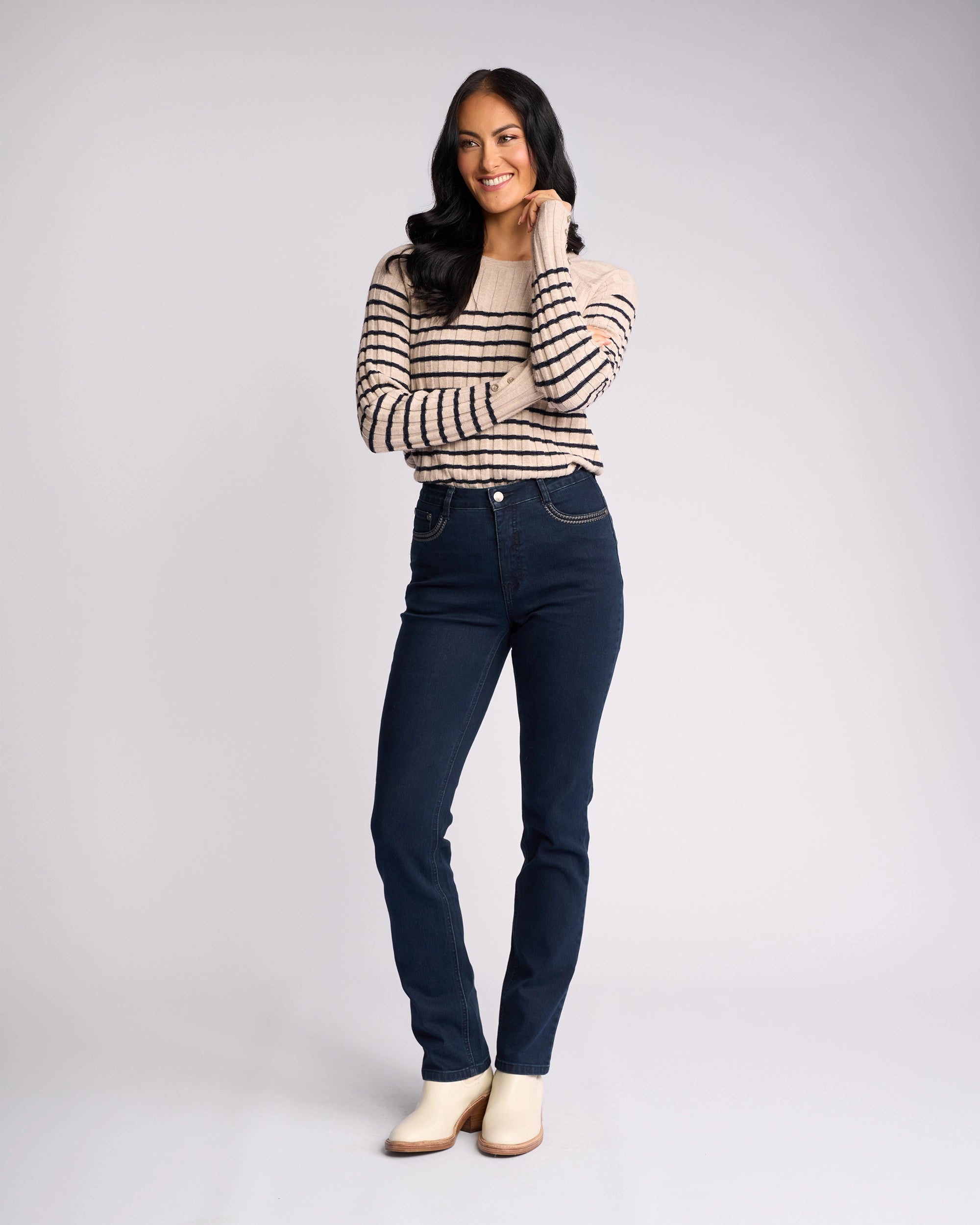 A woman with long dark hair smiles, touching her face, wearing a beige striped sweater, Vassalli’s Urban Jean - Mid Blue high-rise slim-leg jeans, and light ankle boots against a plain light background.