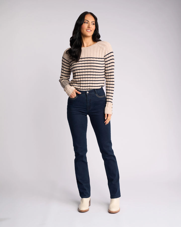 A woman with long dark hair stands in a relaxed pose, wearing a beige and black striped sweater, Vassalli Urban Jean - Mid Blue, and light-colored ankle boots against a plain light background.