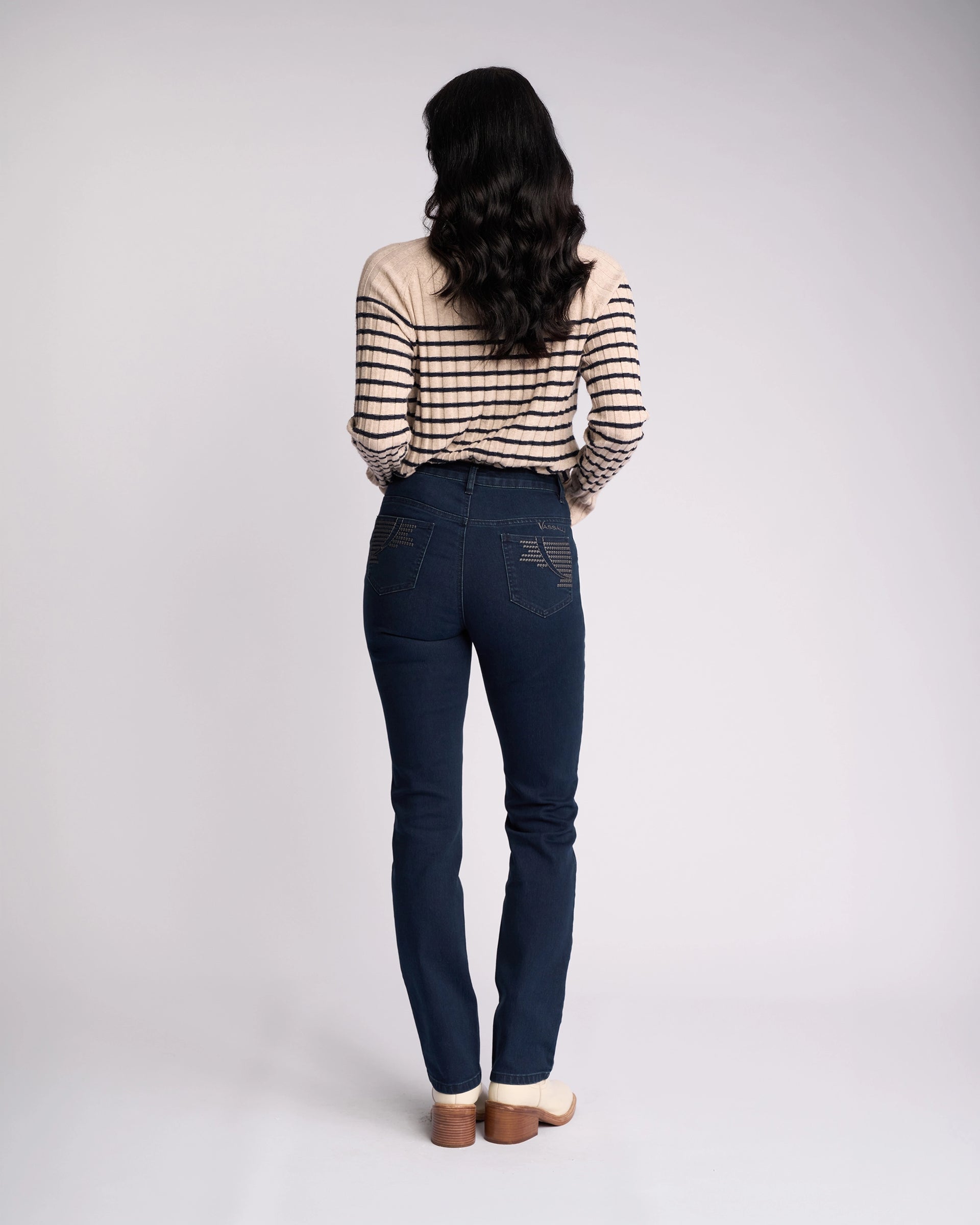 A woman with long dark hair wears a beige and black striped sweater and Vassalli’s Urban Jean - Mid Blue, standing facing away from the camera against a plain light background.