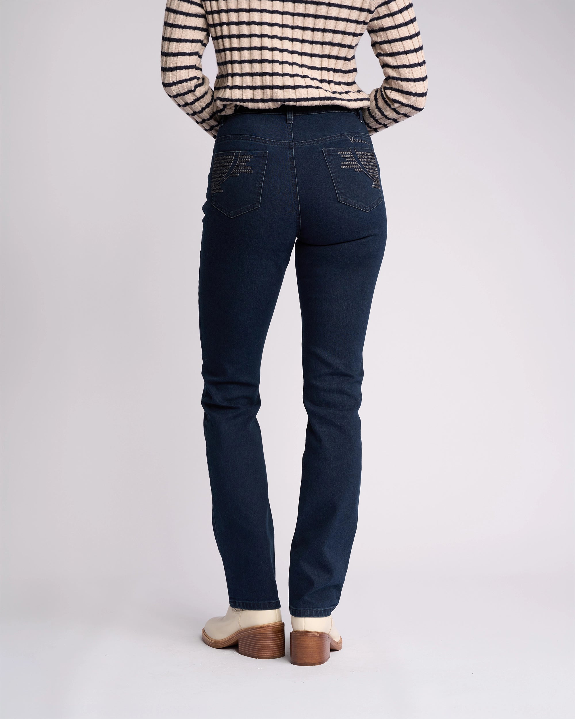 A person stands against a plain background wearing Vassalli Urban Jean - Mid Blue, a long-sleeve beige and black striped top, and cream-colored heeled boots.