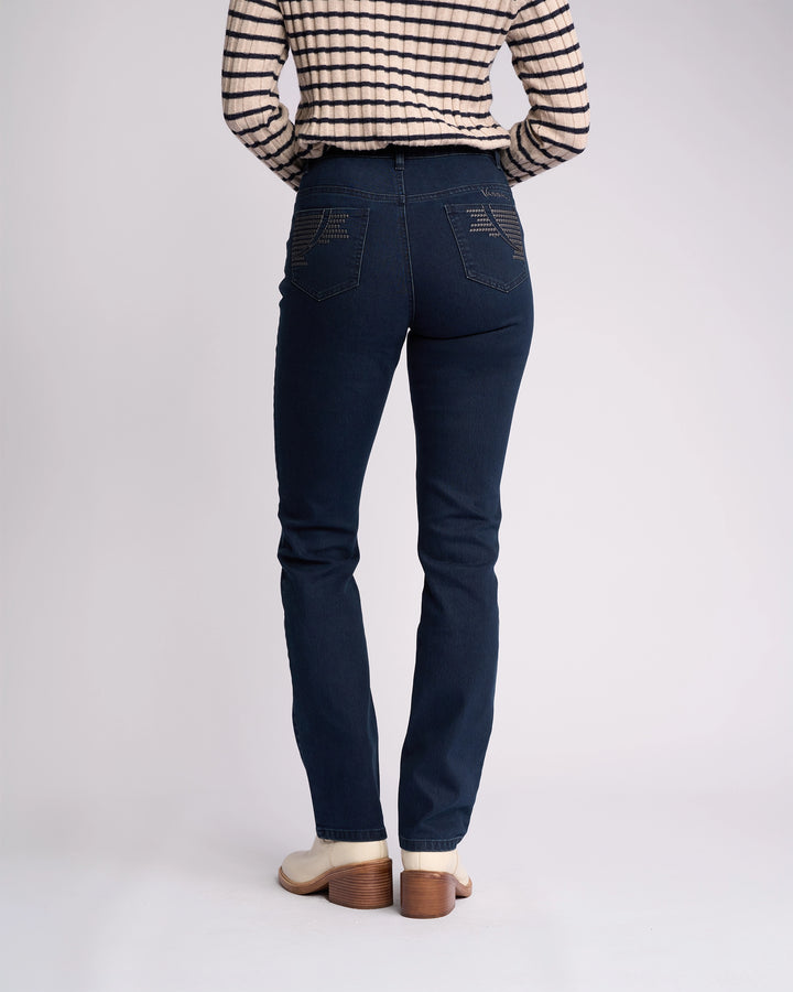 A person stands against a plain background wearing Vassalli Urban Jean - Mid Blue, a long-sleeve beige and black striped top, and cream-colored heeled boots.