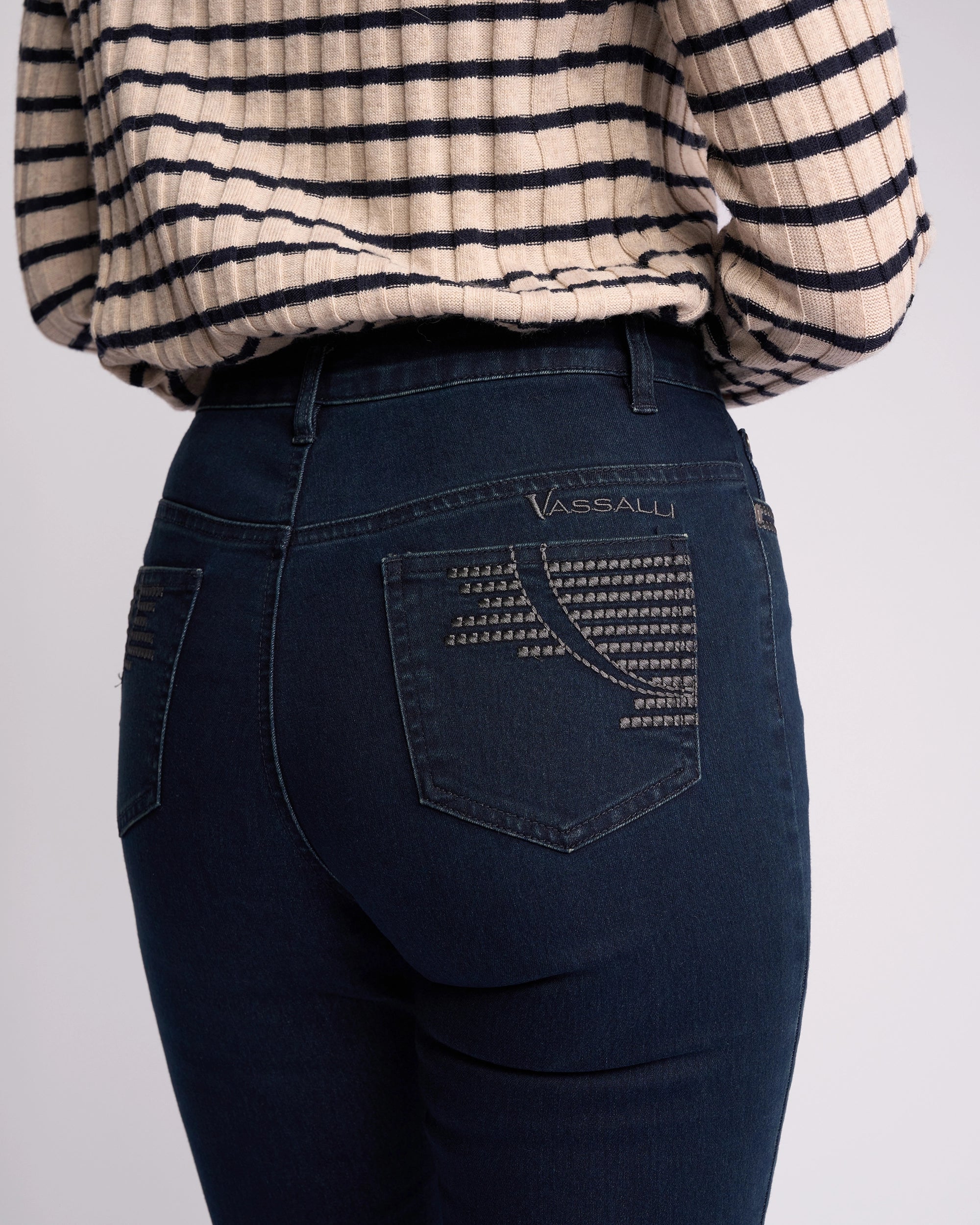 Model wears Vassalli Urban Jean - Mid Blue, a high-rise slim-leg style featuring patterned back pockets and cream/black striped sweater, shown from the back waist down. Vassalli branding appears on the right pocket.